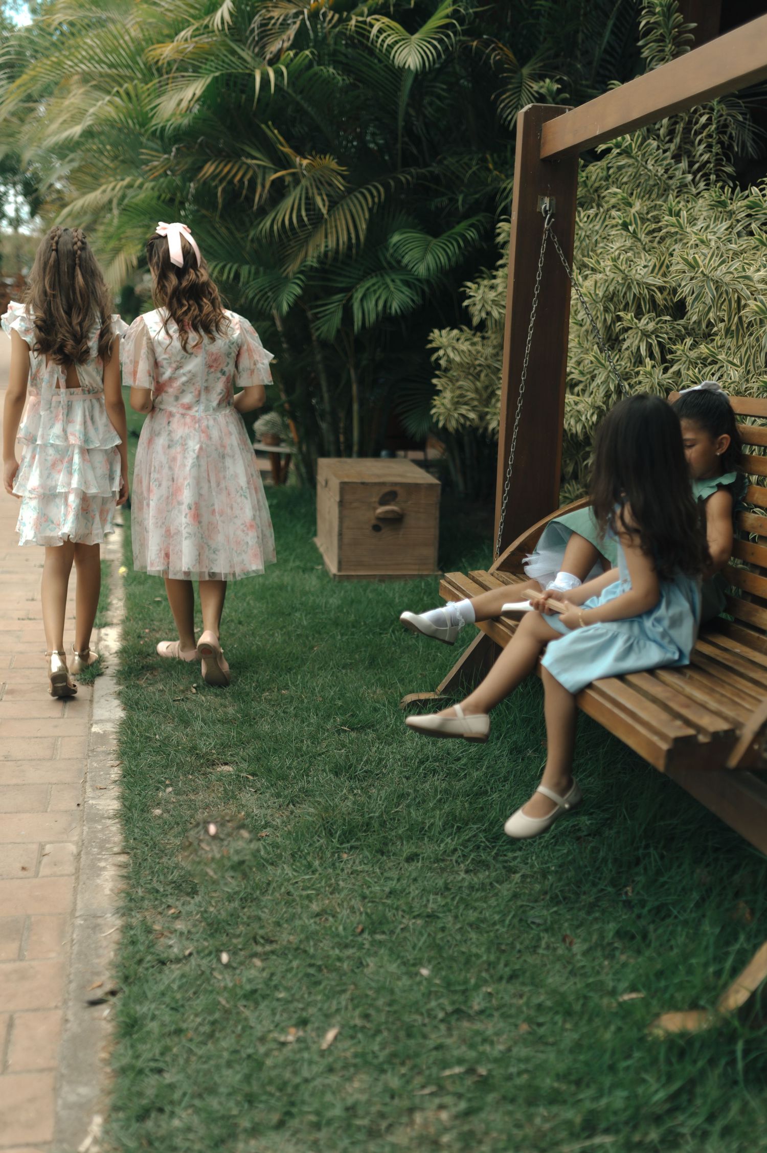 Children in vintage dresses walking and resting on benches in a lush garden path.