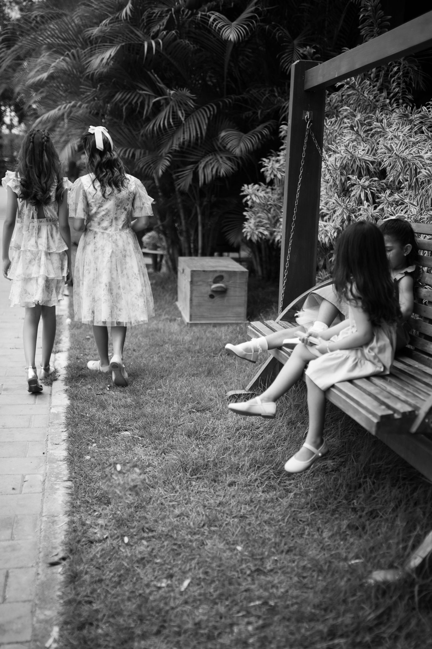 A black and white photo of young people in dresses walking and sitting in a garden setting.