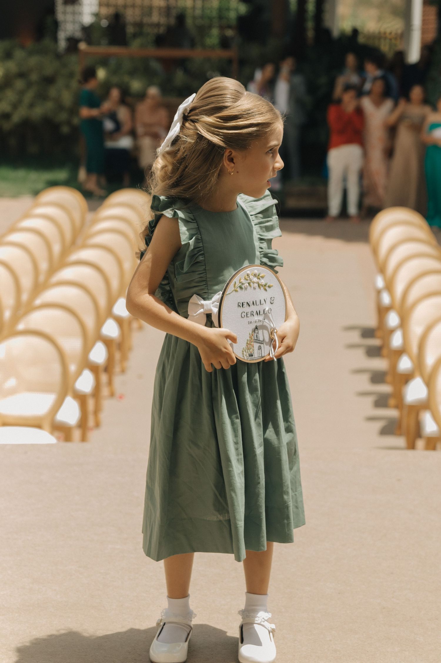 A young attendant in a sage green dress holds a sign while standing in an outdoor wedding aisle.