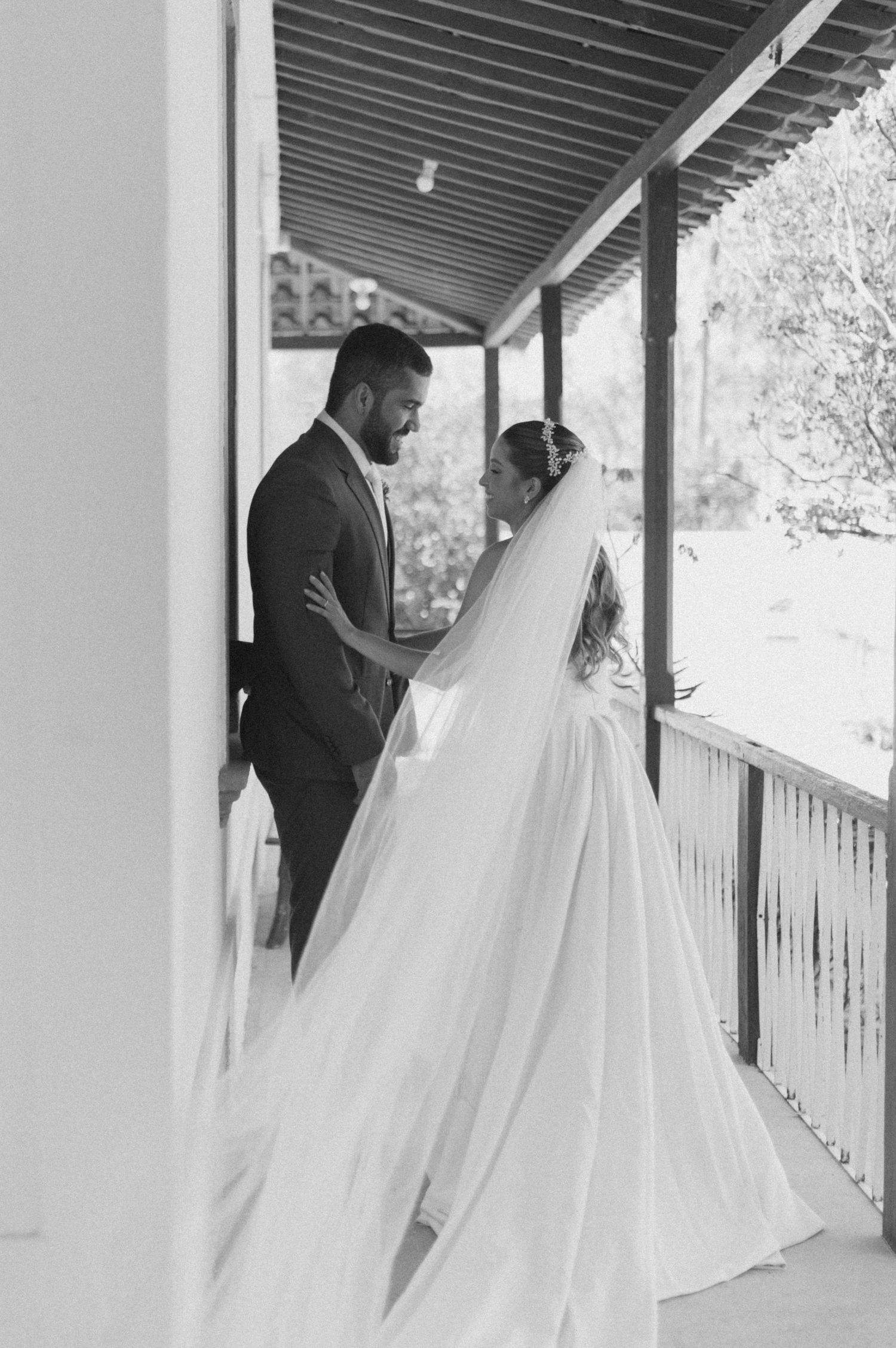 A tender wedding moment between newlyweds sharing an intimate embrace on a covered porch in black and white.