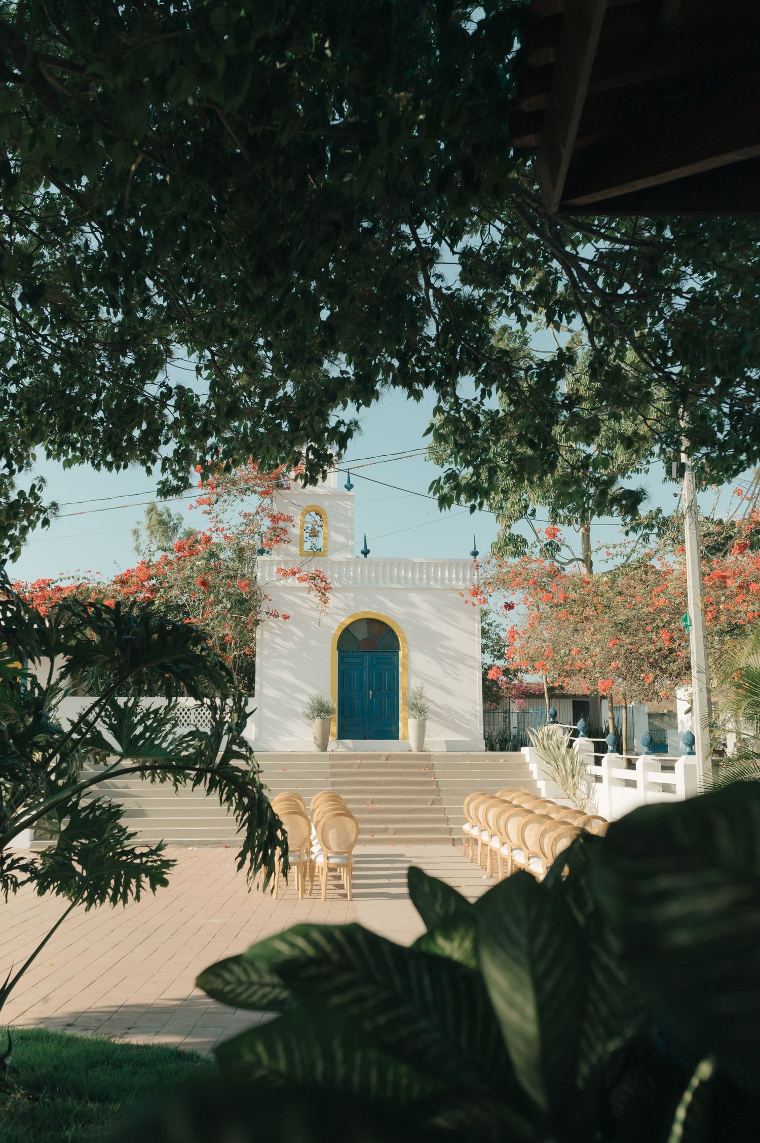 A white church entrance with blue door framed by lush tropical foliage and flowering trees.