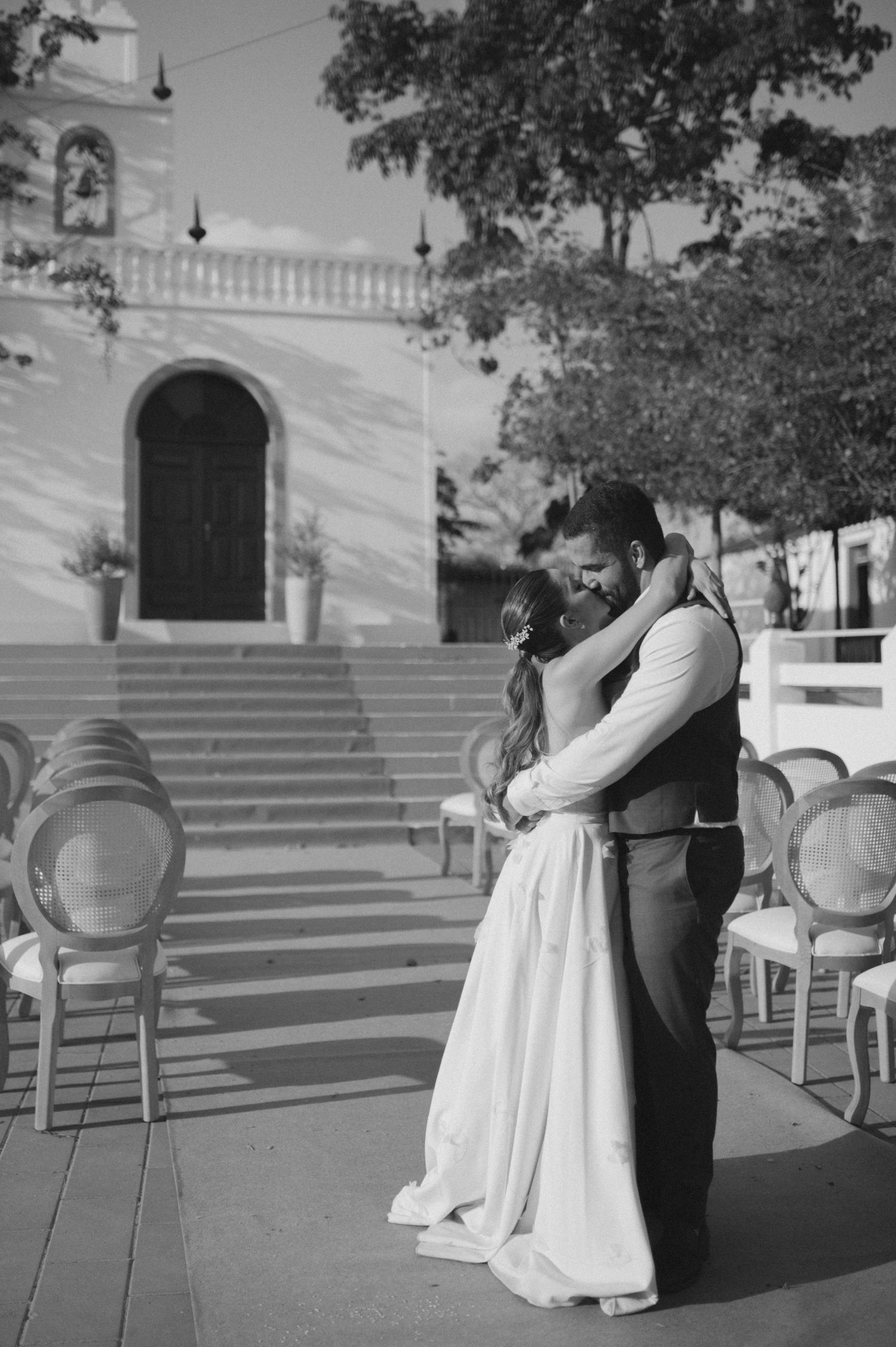 A black and white photo of a wedding couple dancing among empty chairs on a patio.