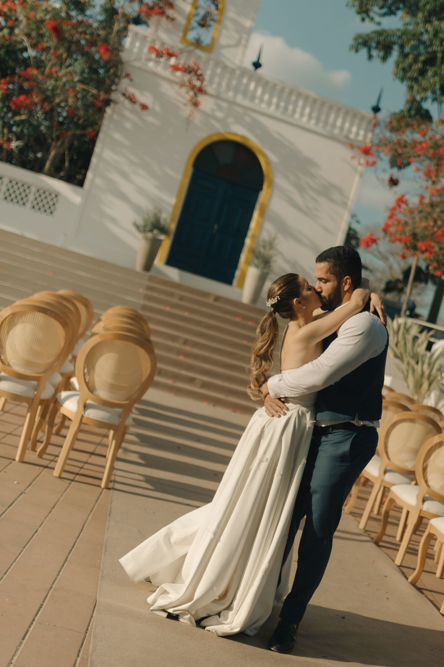 Newlyweds embracing on a sunny patio with elegant chairs and blooming trees.