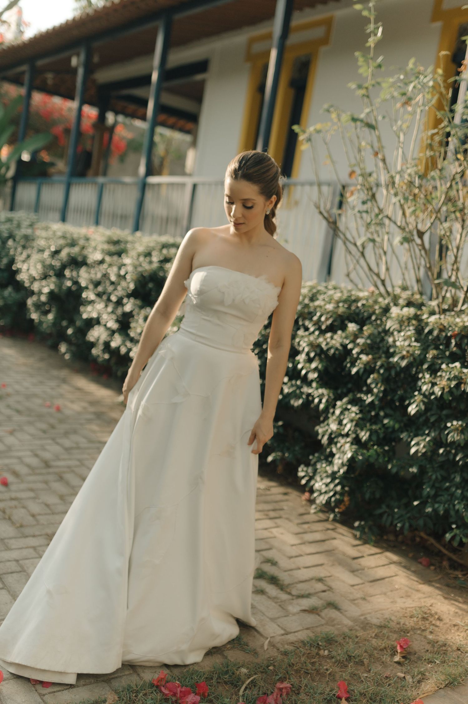 Elegant bride in a minimalist white dress stands on a brick path surrounded by flowers.