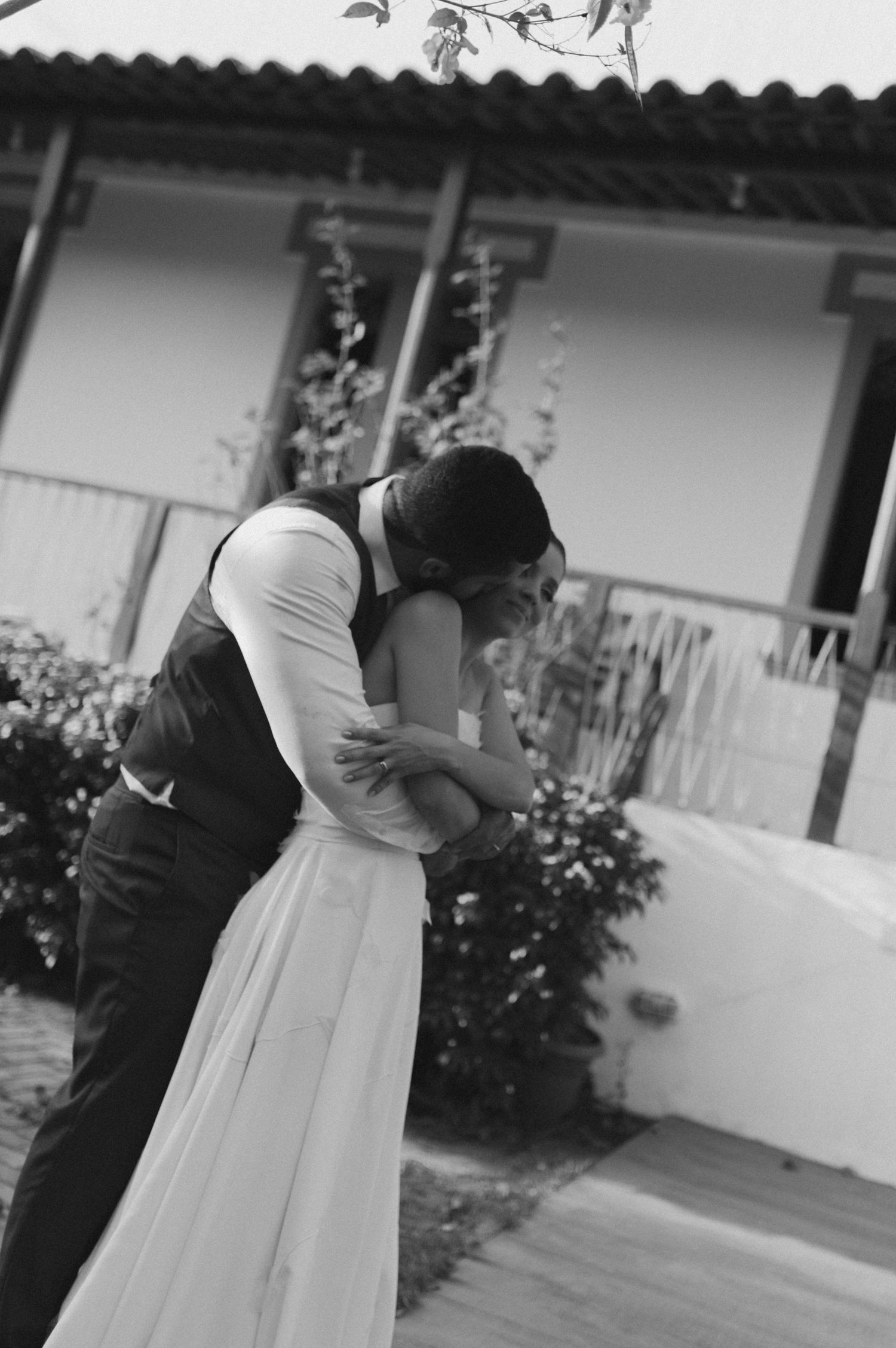 A black and white photo of a romantic embrace between newlyweds in front of a white building.