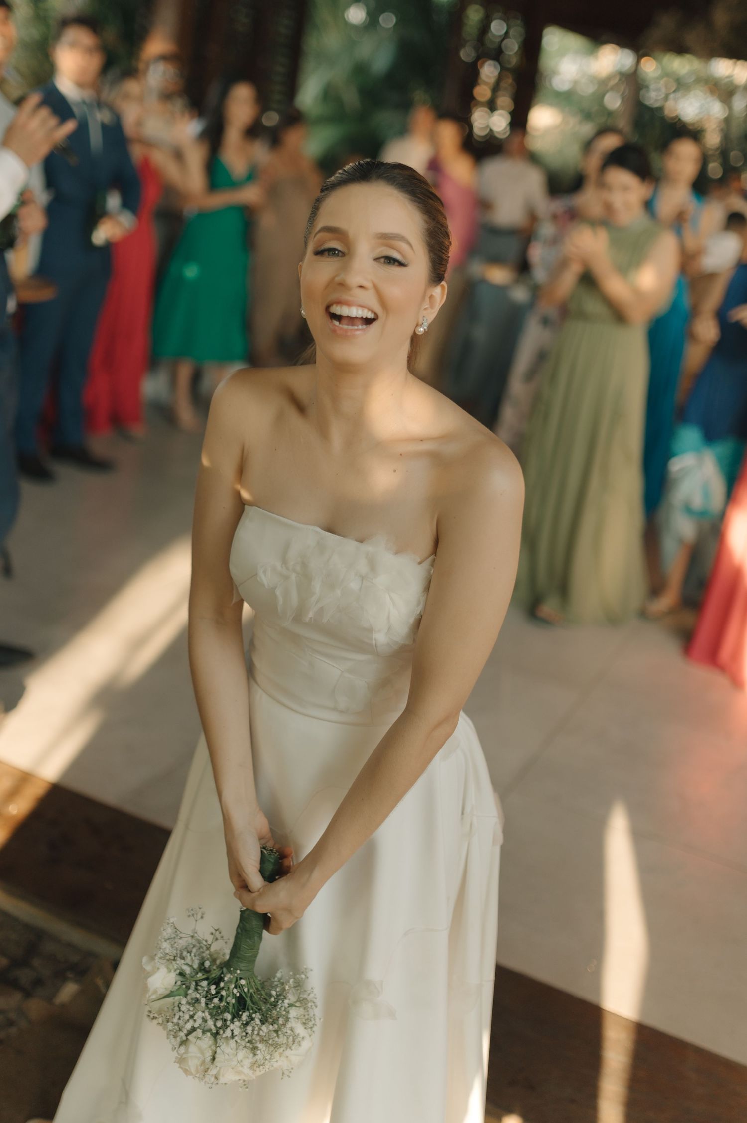 A bride in a white strapless dress holding a bouquet smiles joyfully during her wedding reception.