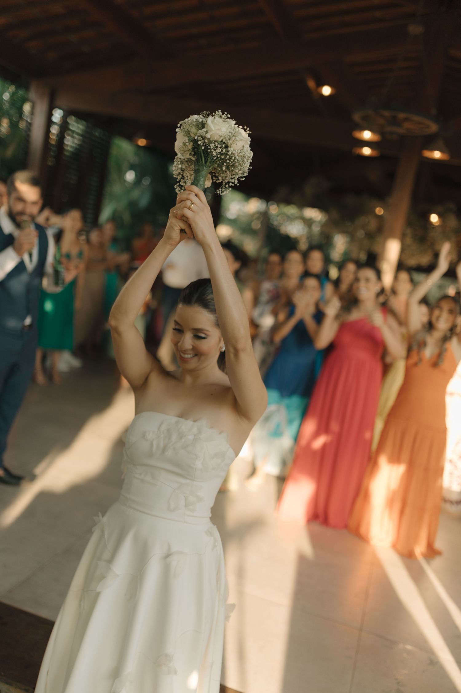 A bride in white holds up her bouquet while celebrating with wedding guests in colorful dresses.