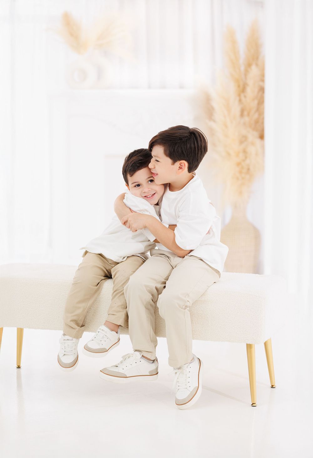 Young boy in white shirt and khaki pants poses playfully on white chairs in a bright studio setting.