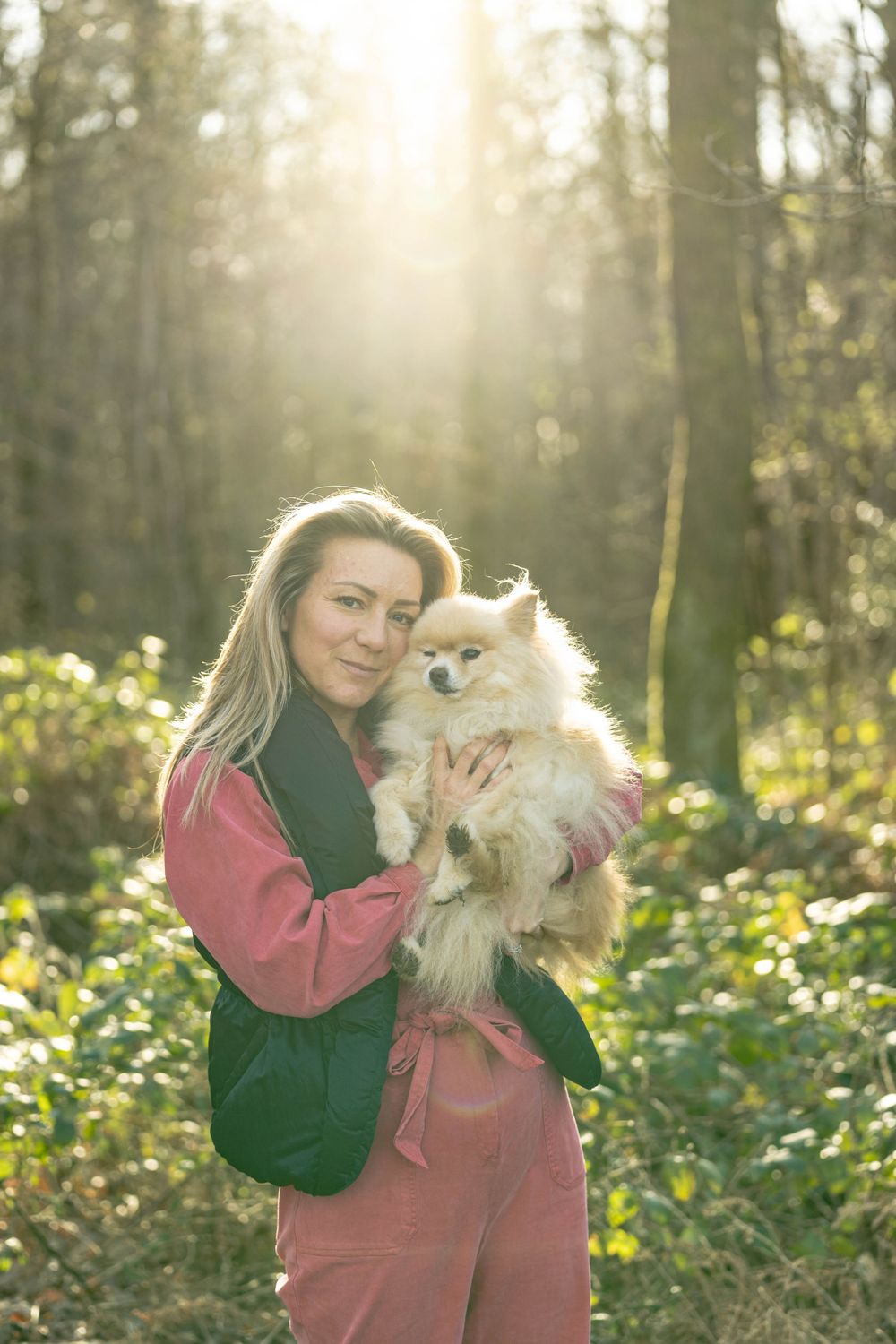 personal branding photography of a woman holding her dog in the forest wearing a red jumpsuit with long hair. the sunlight through trees