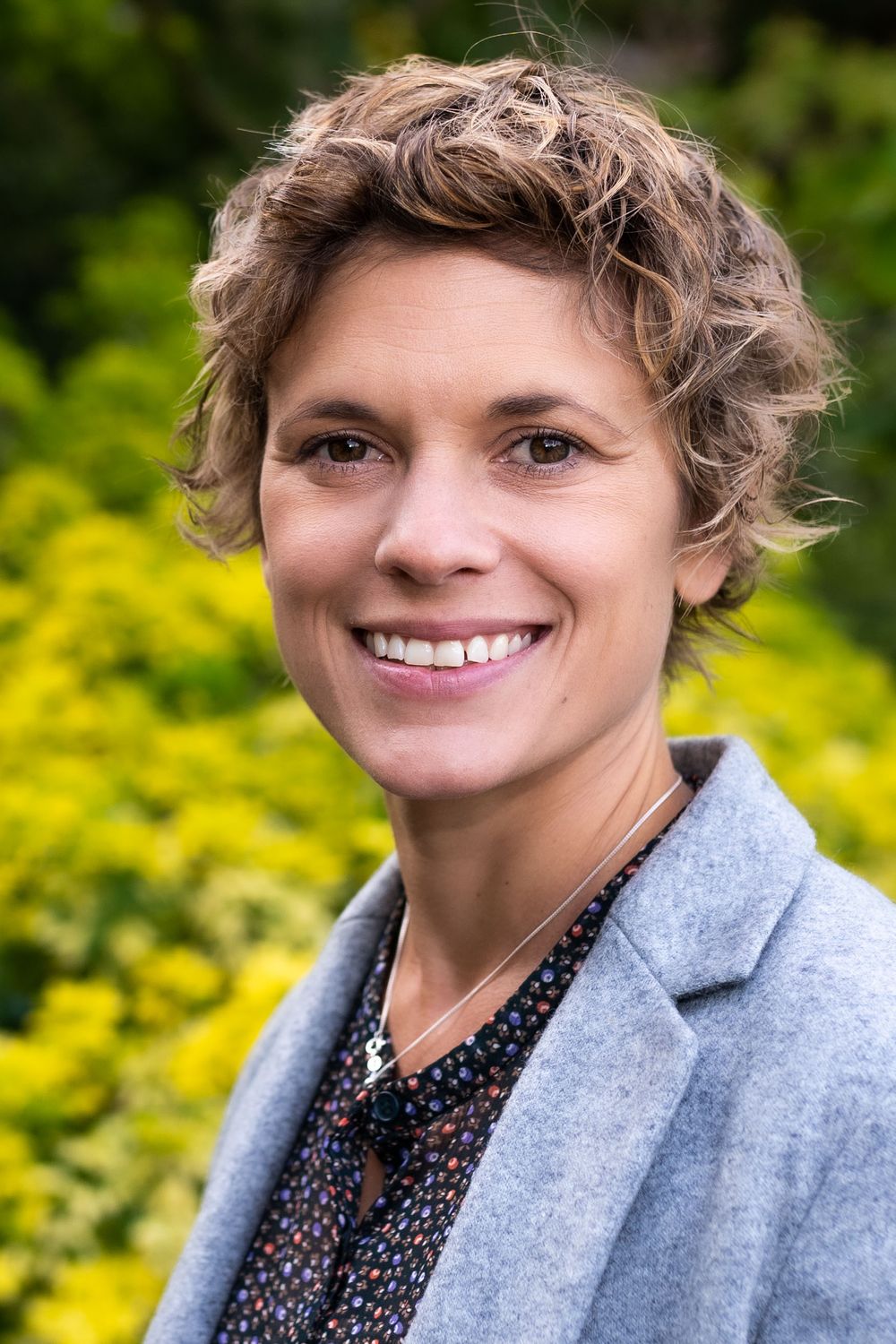 Headshots Brighton and sussex a woman smile to camera in a garden she wears a grey jacket and hasher curly hair