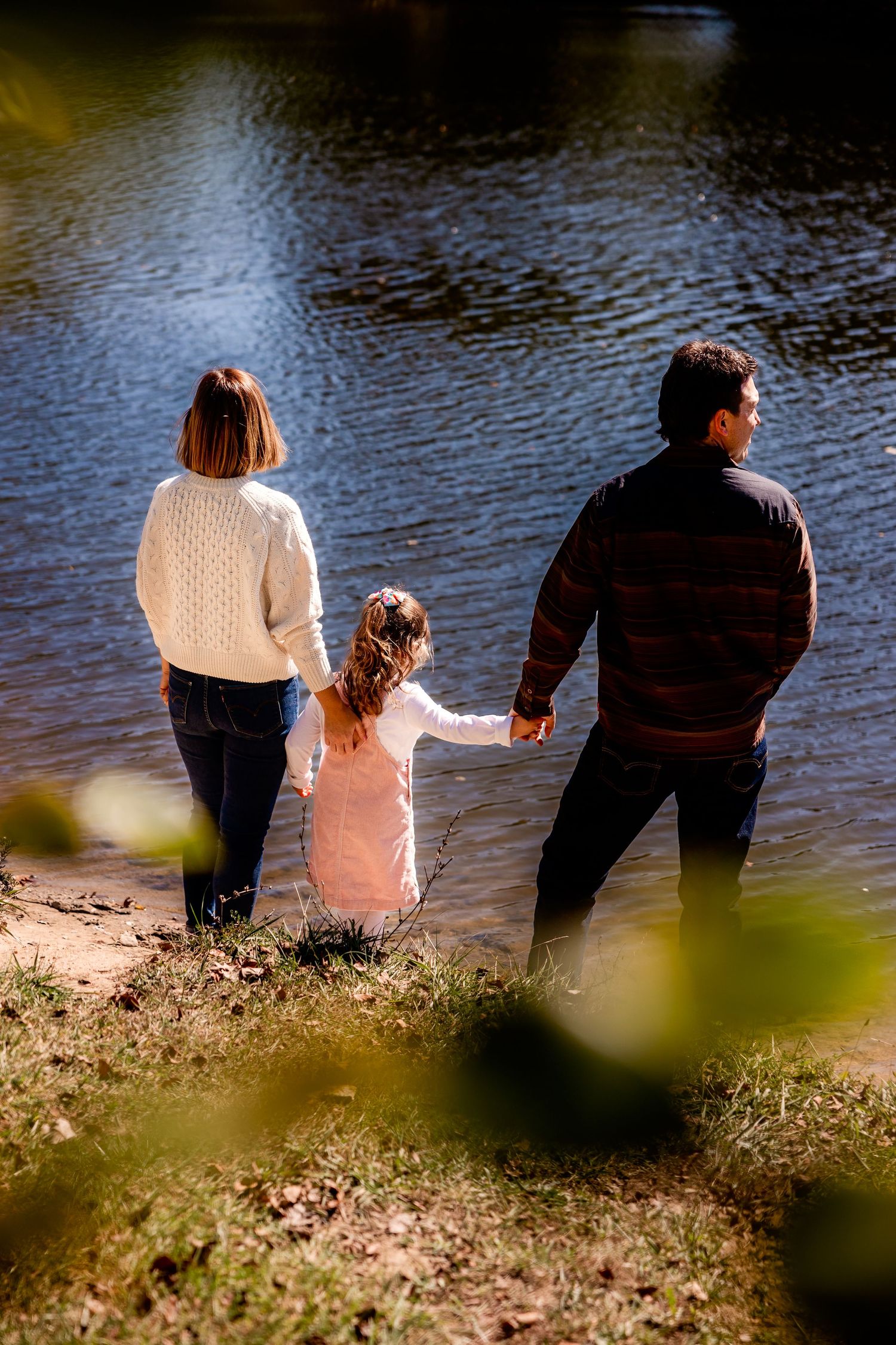Three people stand holding hands at the edge of a lake on a sunny day, viewing the calm water together. Photo by Rachel Martinez Photography