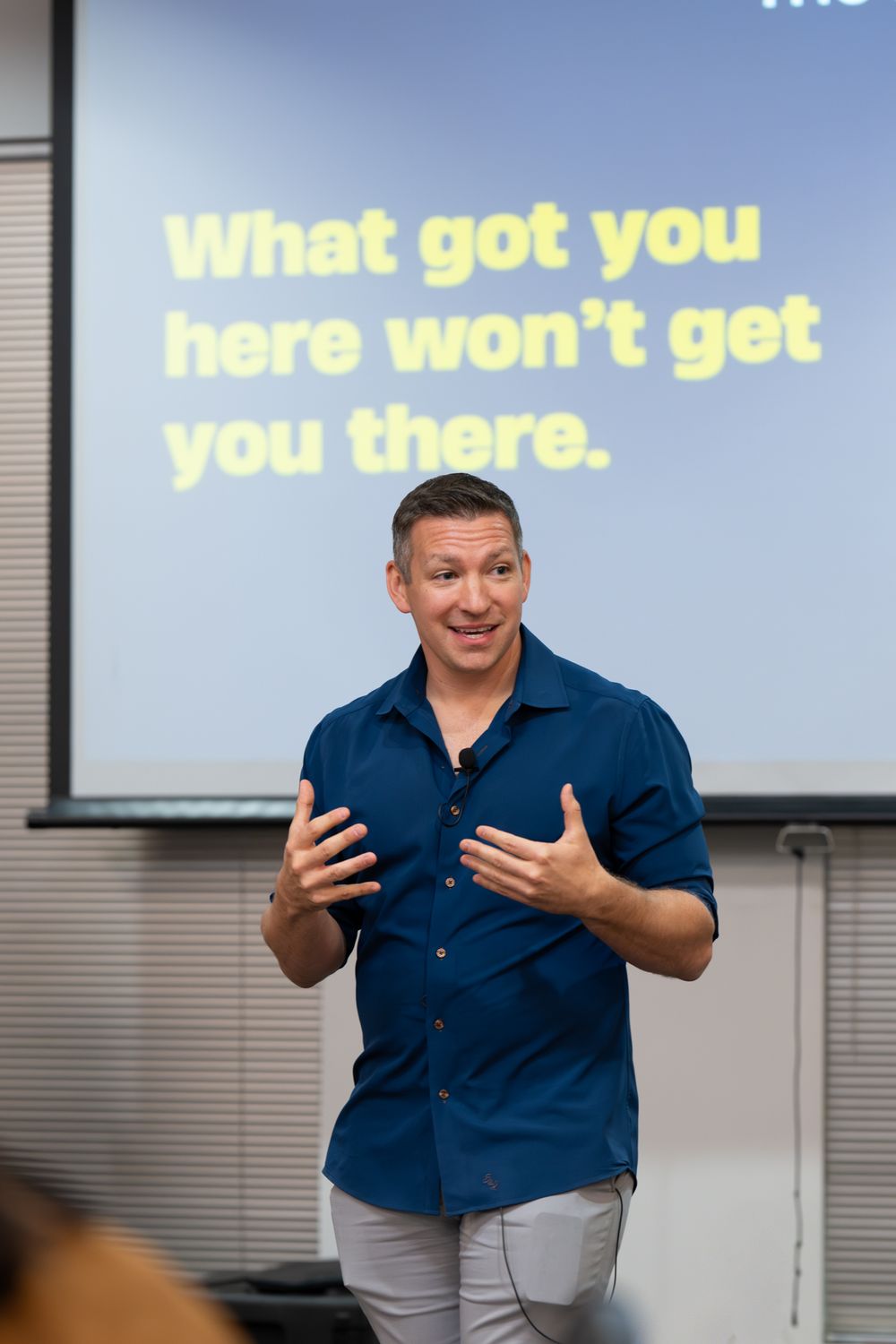 A presenter in a navy blue shirt gestures animatedly while speaking in front of a projection screen.