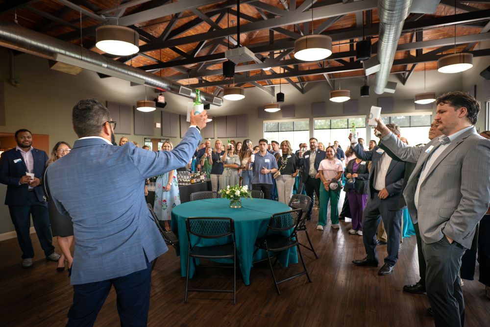 Guests raise glasses in a toast at an elegant indoor event with teal table centerpieces and exposed ceiling beams.