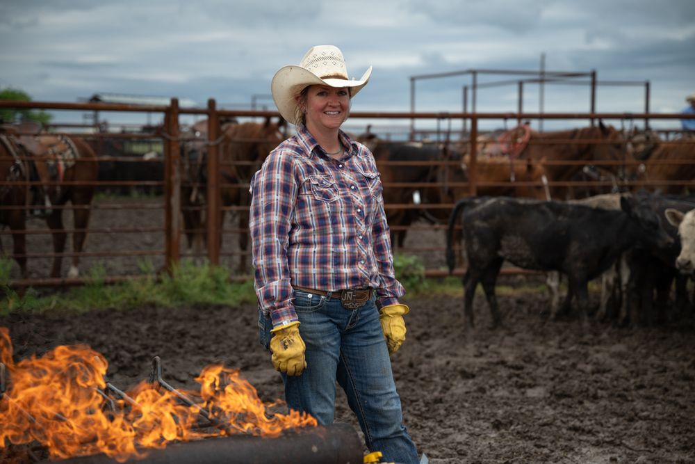 A cowboy wearing yellow gloves works with cattle in a dusty ranch corral as flames rise in the background.