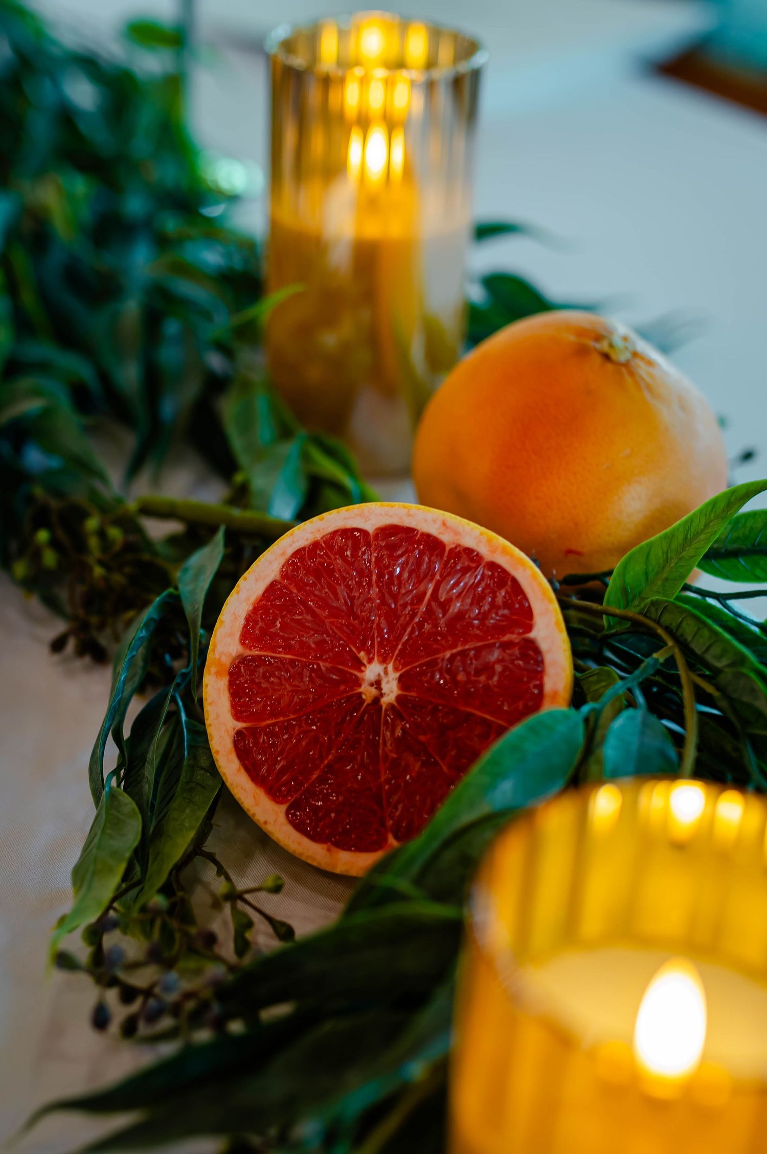 Halved red grapefruit and whole orange arranged with green leaves and glowing candles in glass holders.