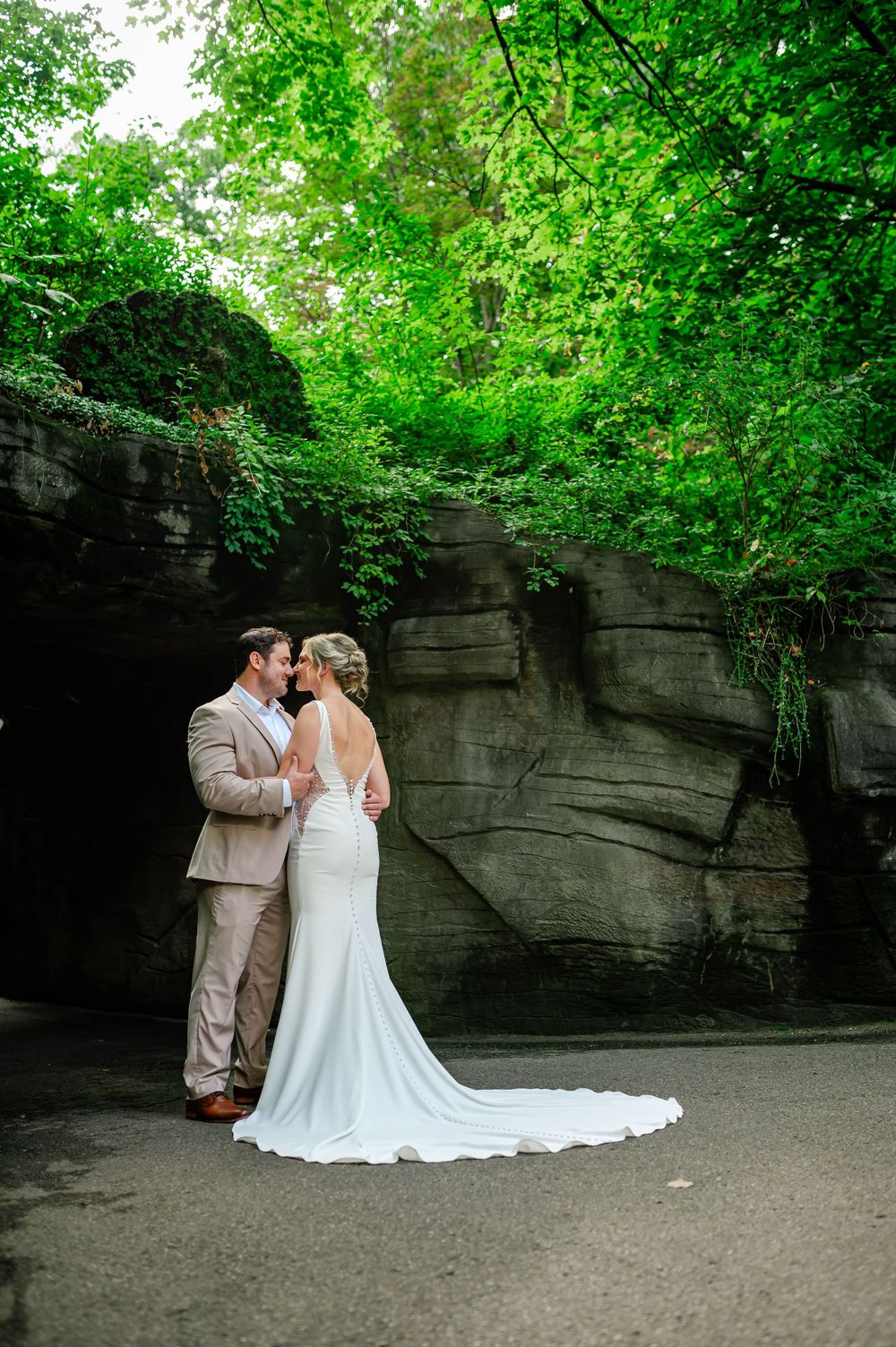 Two people share a romantic moment in white wedding attire against a backdrop of rocky cliffs and lush greenery.