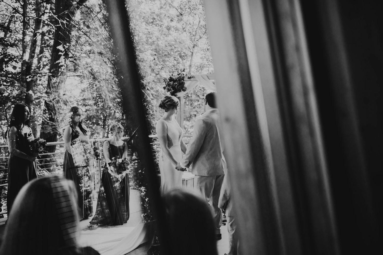 A black and white outdoor wedding ceremony scene framed through curtains showing a couple at the altar.