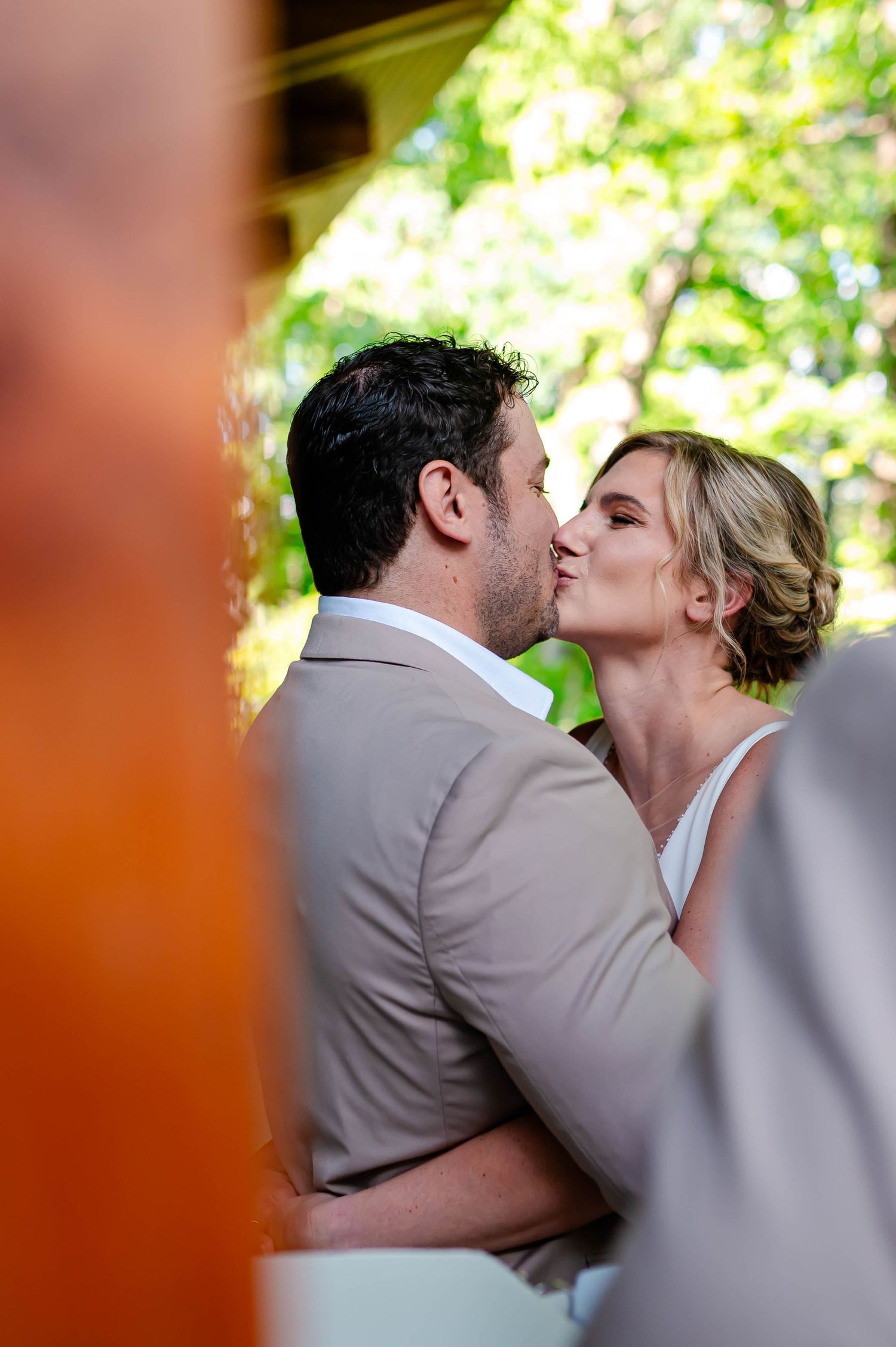 A romantic wedding kiss shared between a bride in white and groom in beige suit in an outdoor garden setting.