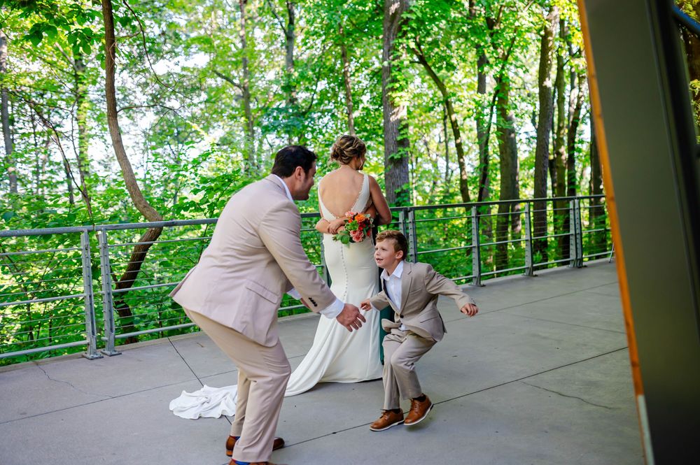 A wedding party in light pink suits and white dress gather on a covered deck surrounded by lush green forest scenery.