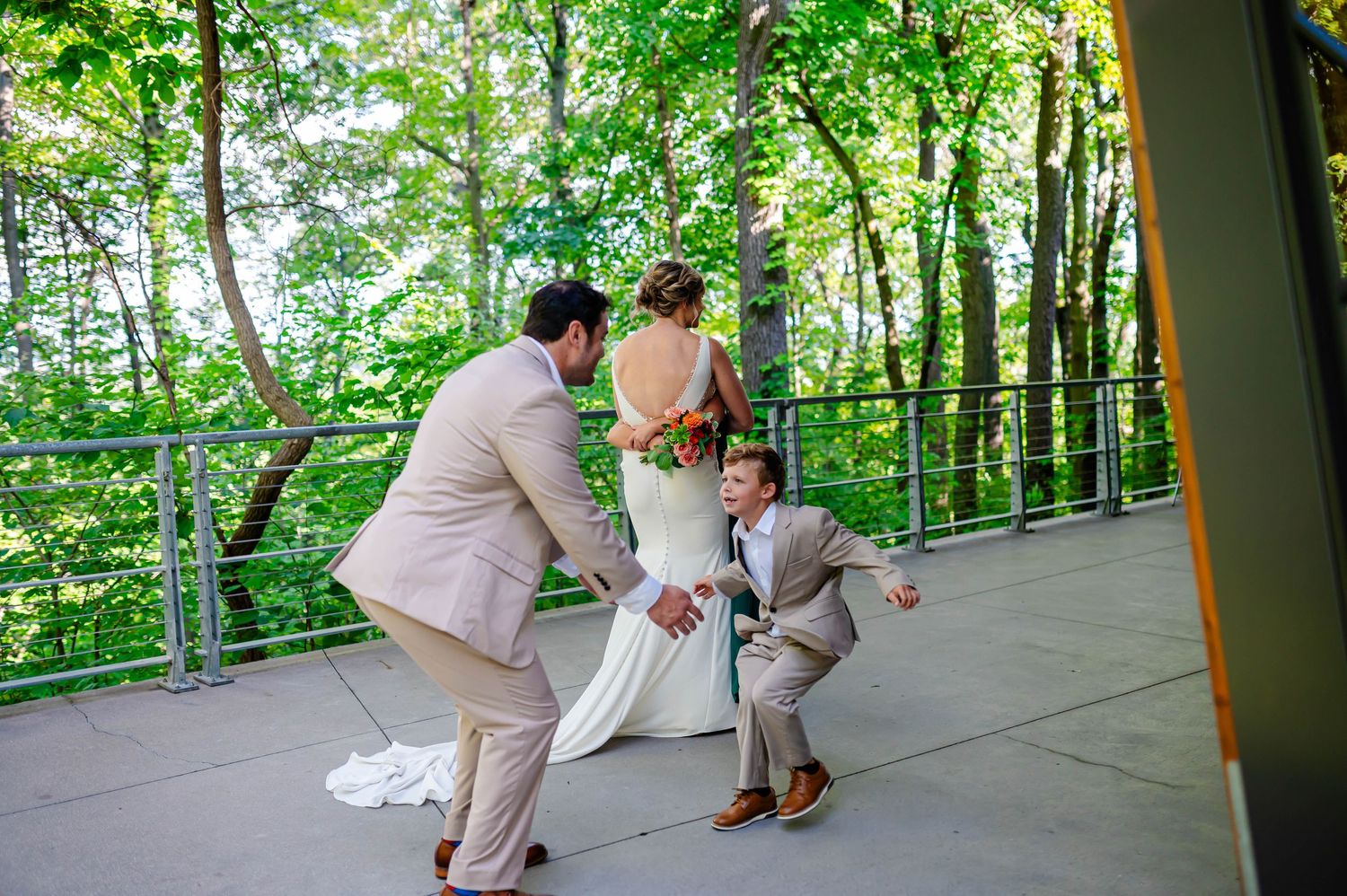 A wedding party in light pink suits and white dress gather on a covered deck surrounded by lush green forest scenery.