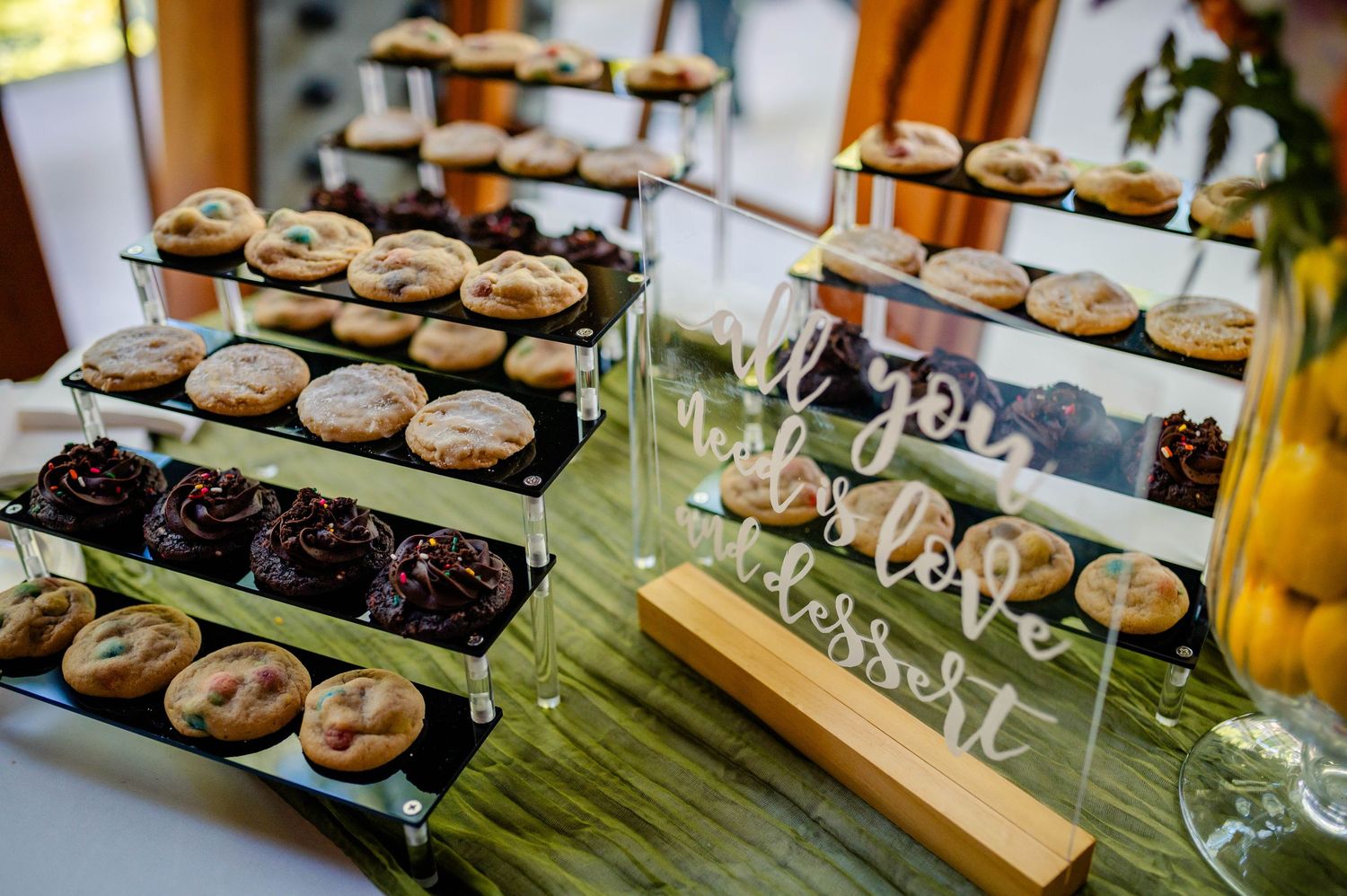 Tiered display of freshly baked chocolate chip and dark chocolate cookies arranged on wooden stands with wine glasses.