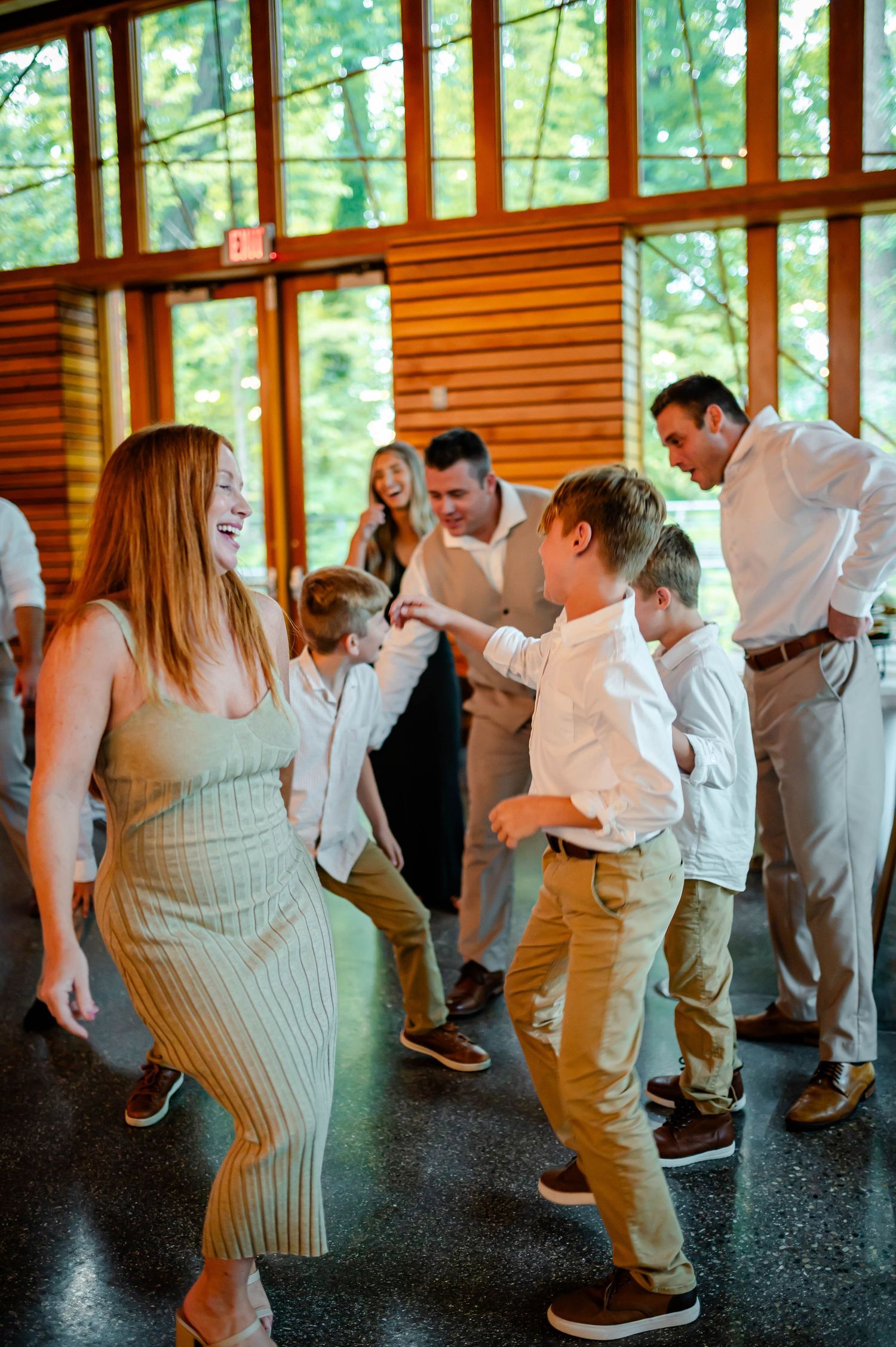 Wedding guests dancing energetically in a bright room with wood paneled walls and large windows.