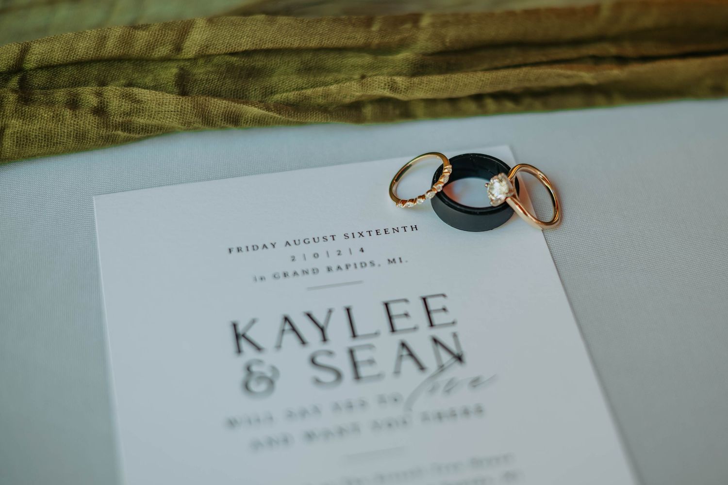 Wedding invitation card and rings resting on a white surface with soft background lighting.