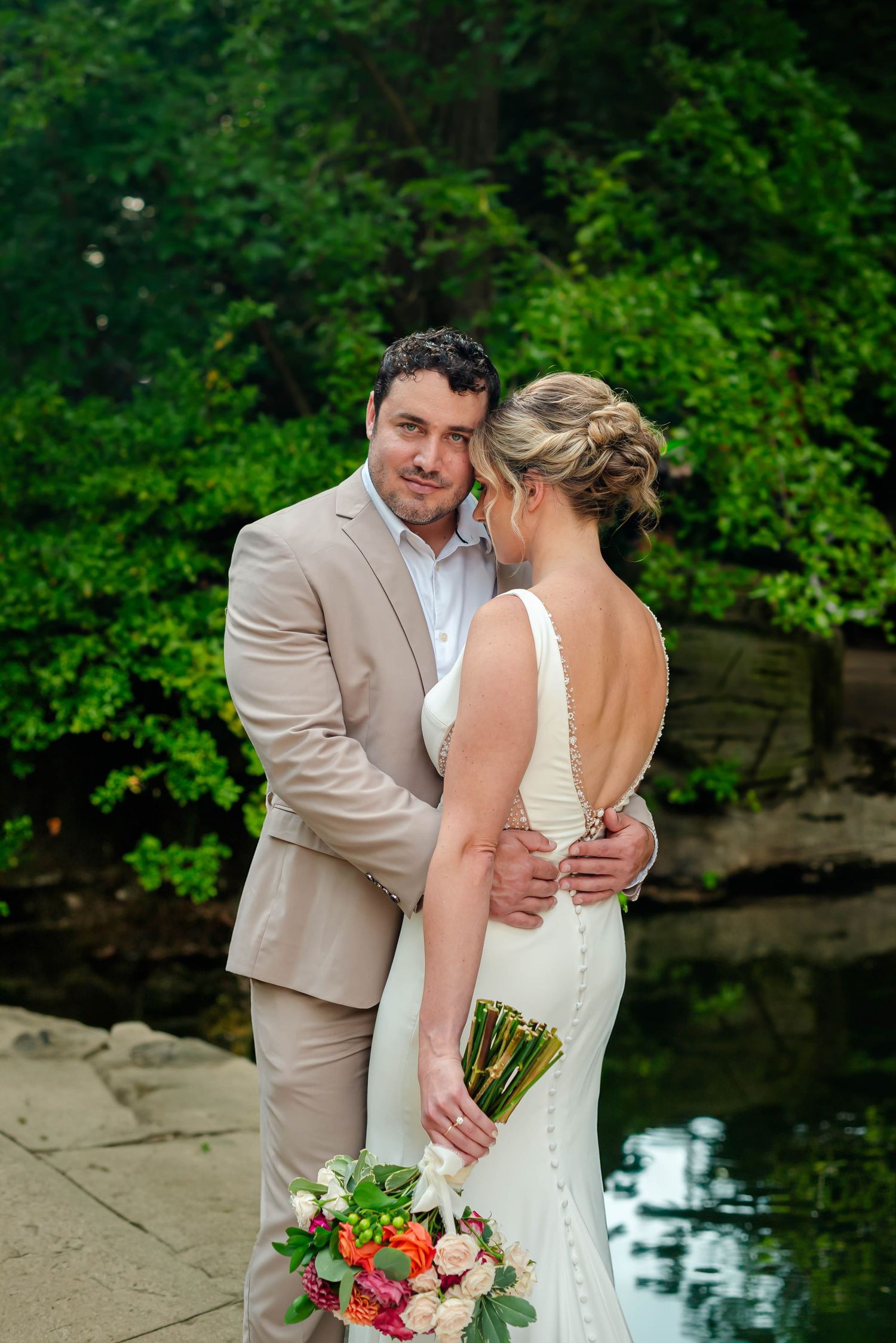 Newlyweds share tender moments by a tranquil pond surrounded by lush greenery during their wedding photoshoot.