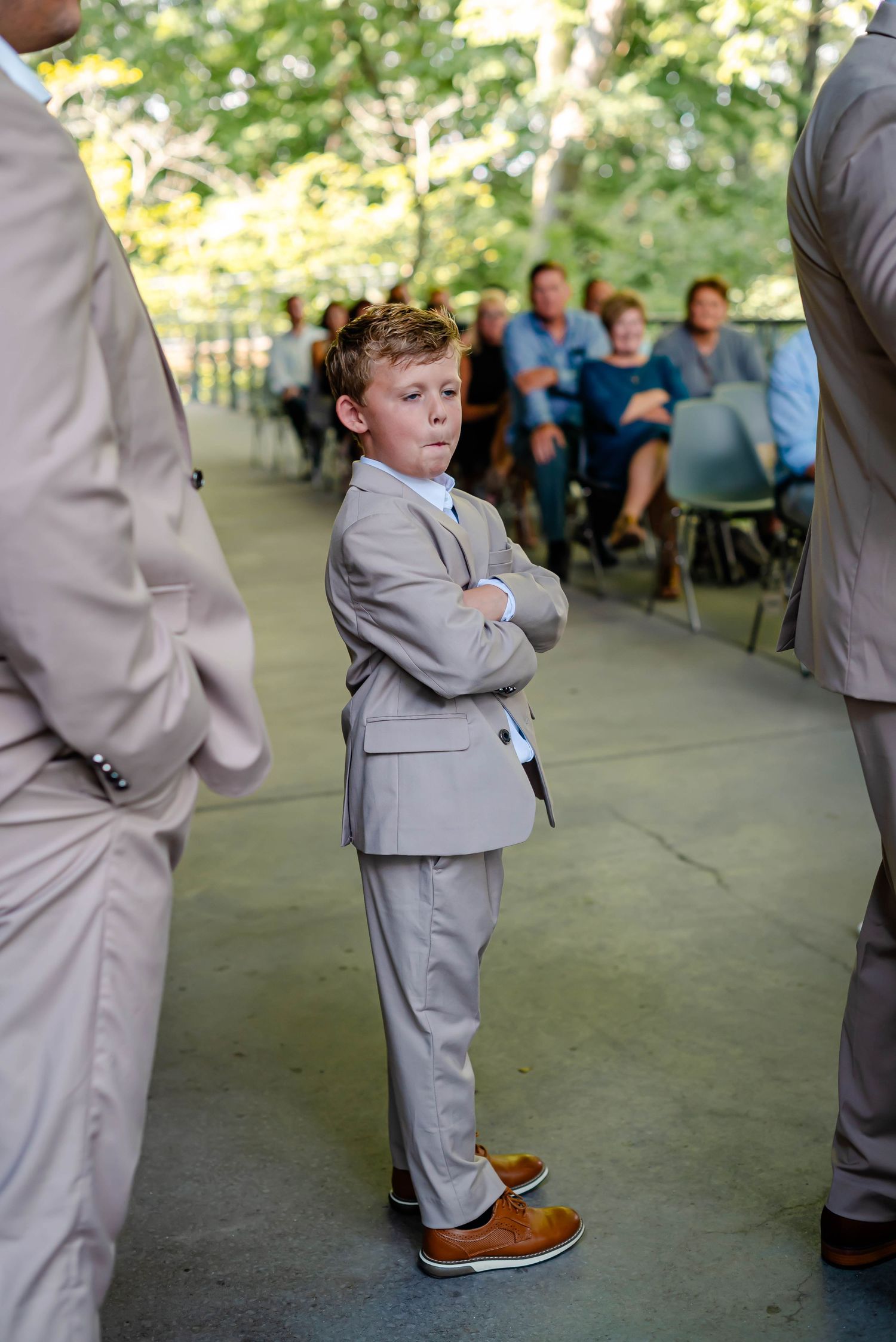 Young boys wearing grey suits stand in line at a formal event with outdoor backdrop.