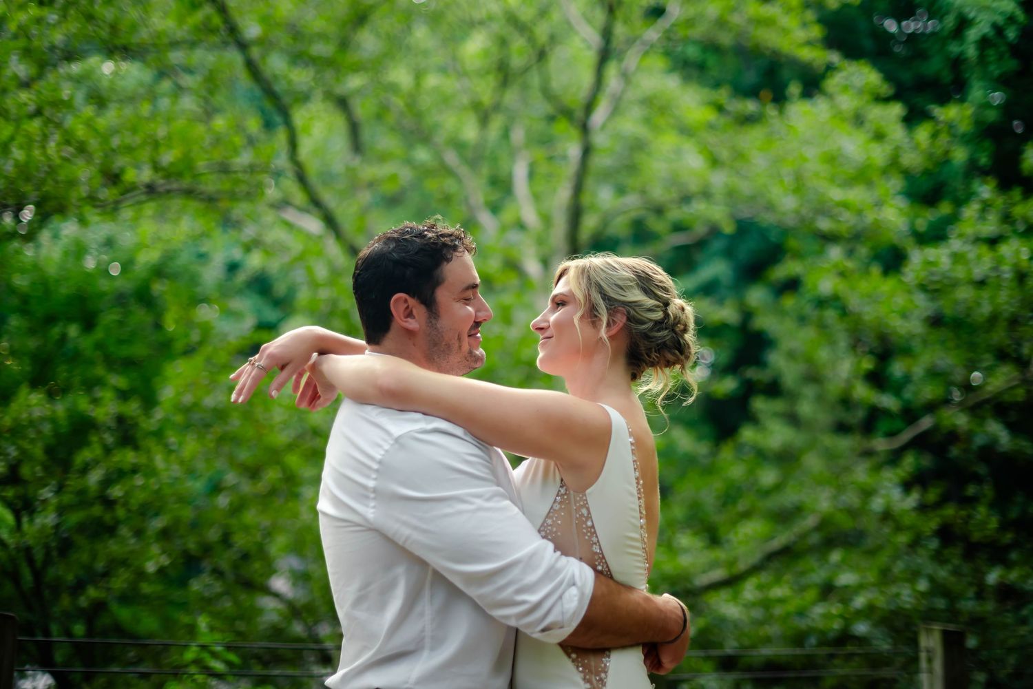 A couple in white embraces during their outdoor wedding photoshoot with lush greenery in the background.