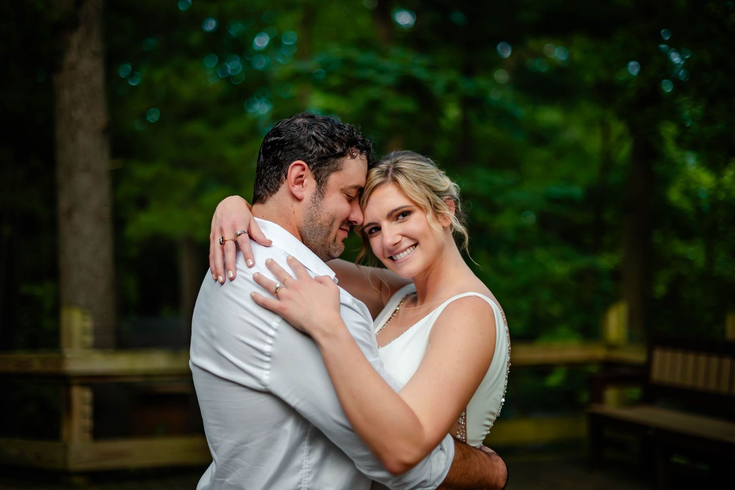 A couple in white attire shares tender moments during an outdoor photoshoot in a natural wooded setting.