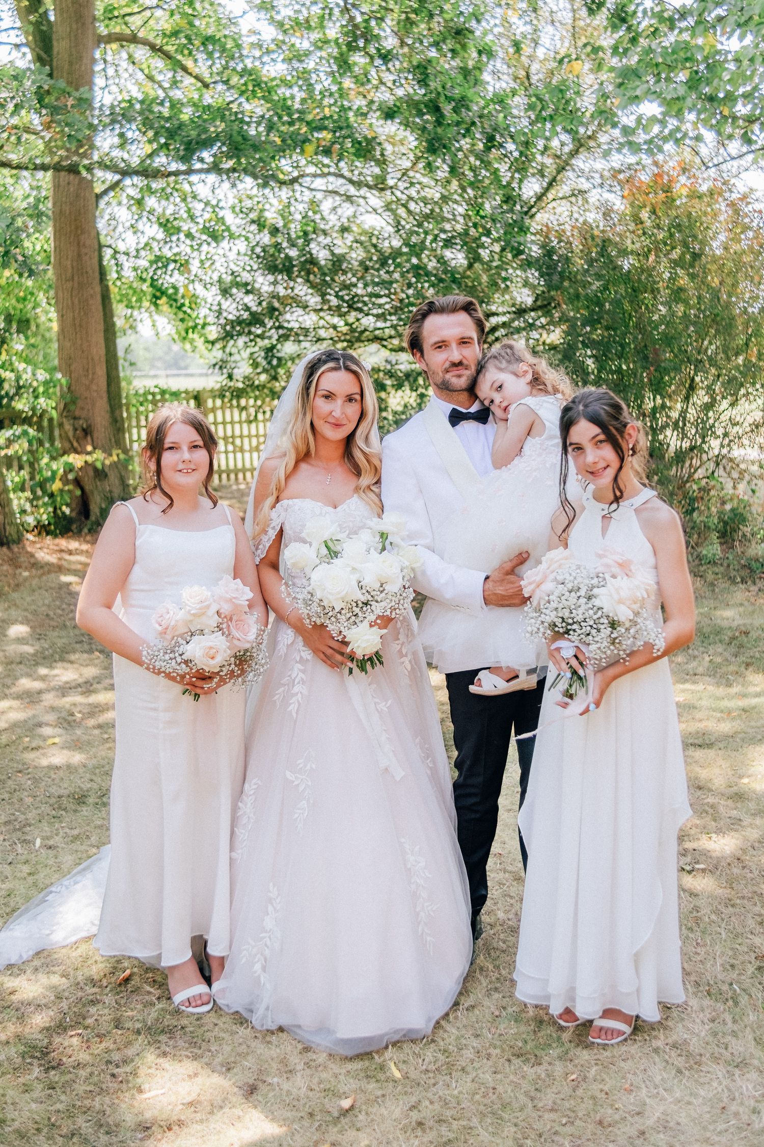 A couple in wedding attire embraces on a path with white flowers and rustic fence in a romantic outdoor setting.