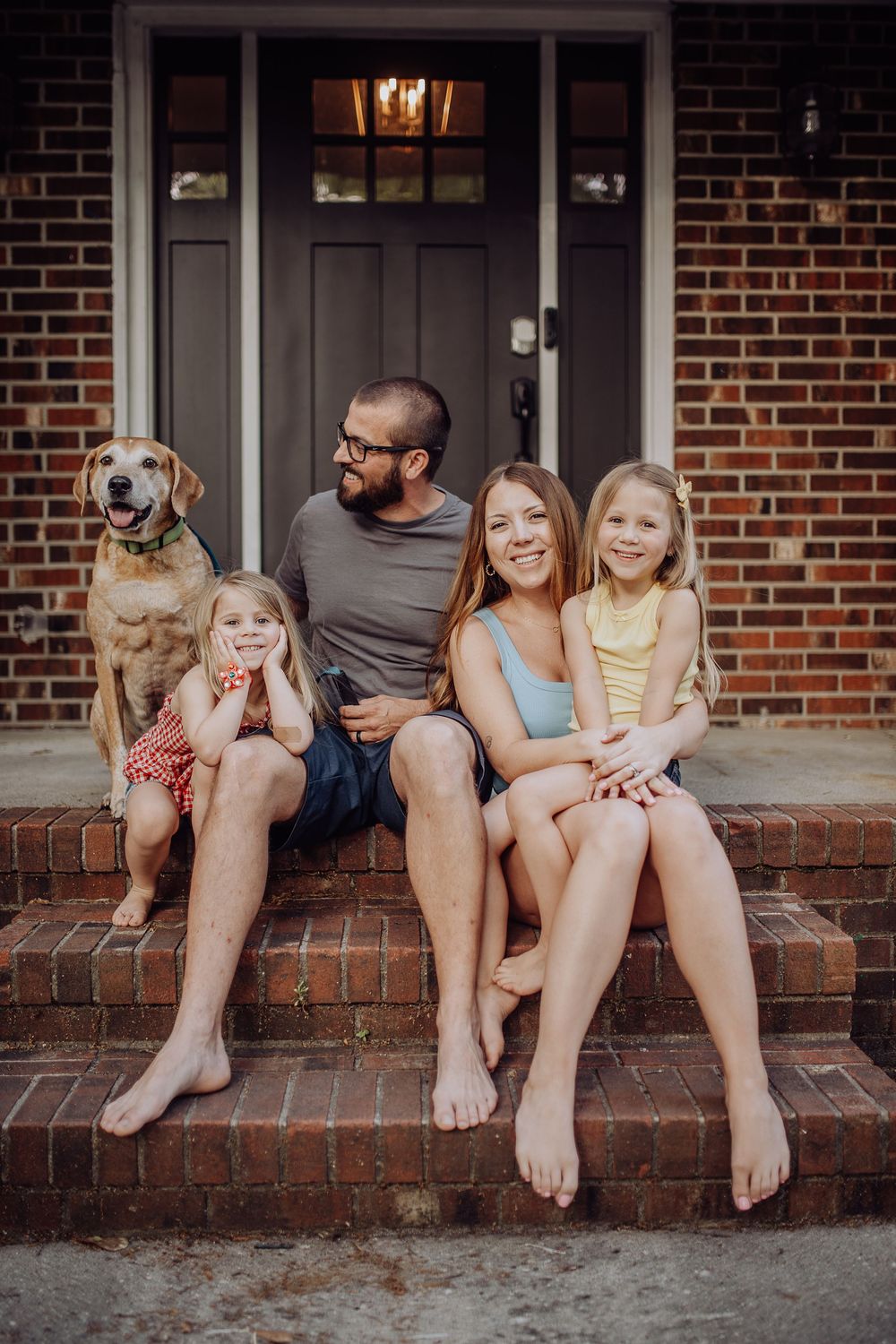Family of four with dog sitting on brick front porch steps of home.