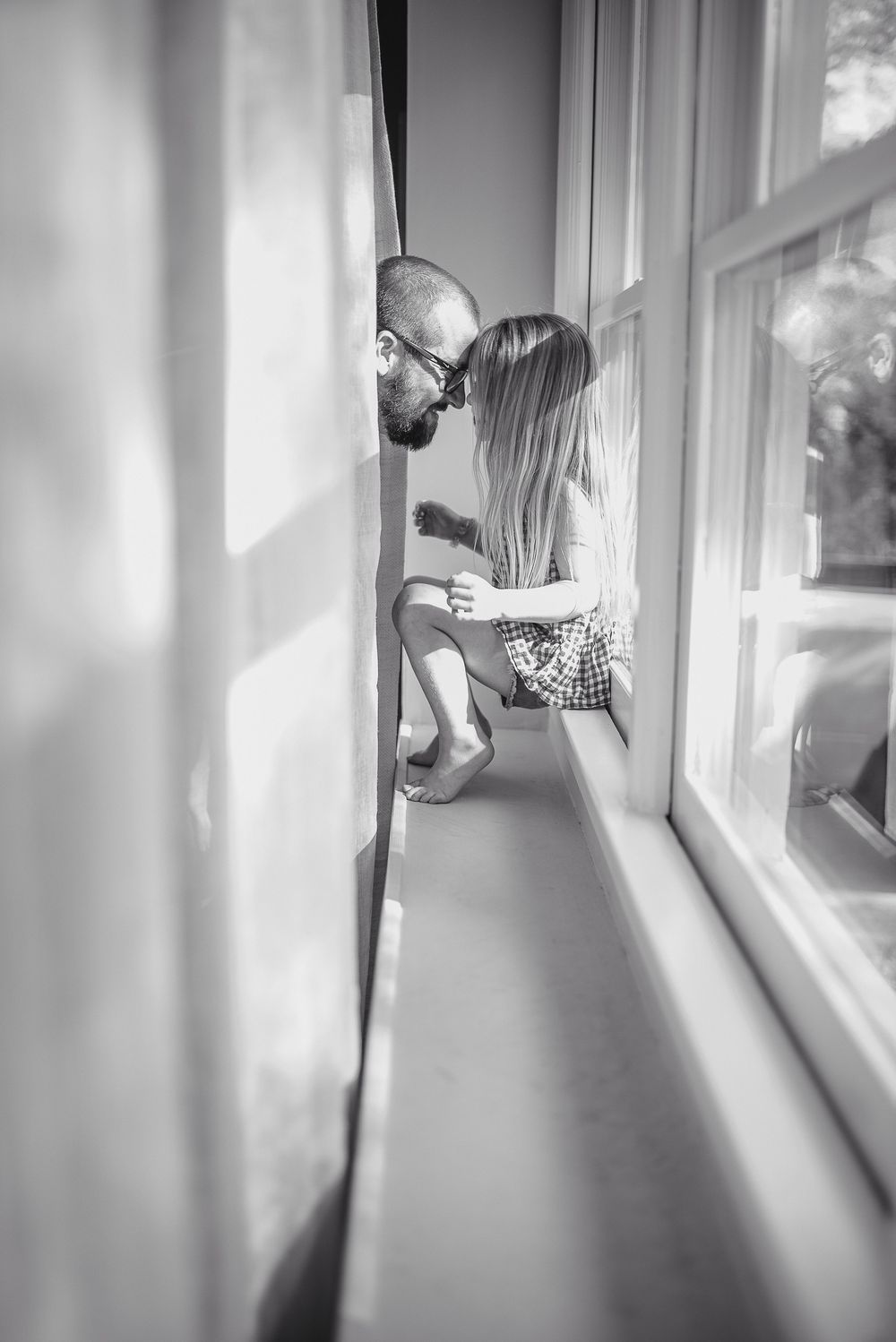 Black and white photo of a father and young daughter sharing a tender moment together on a sunlit window ledge.