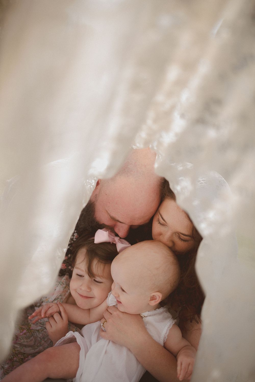 A bald father and dark-haired mother cuddle their two young daughters, viewed through soft white fabric.
