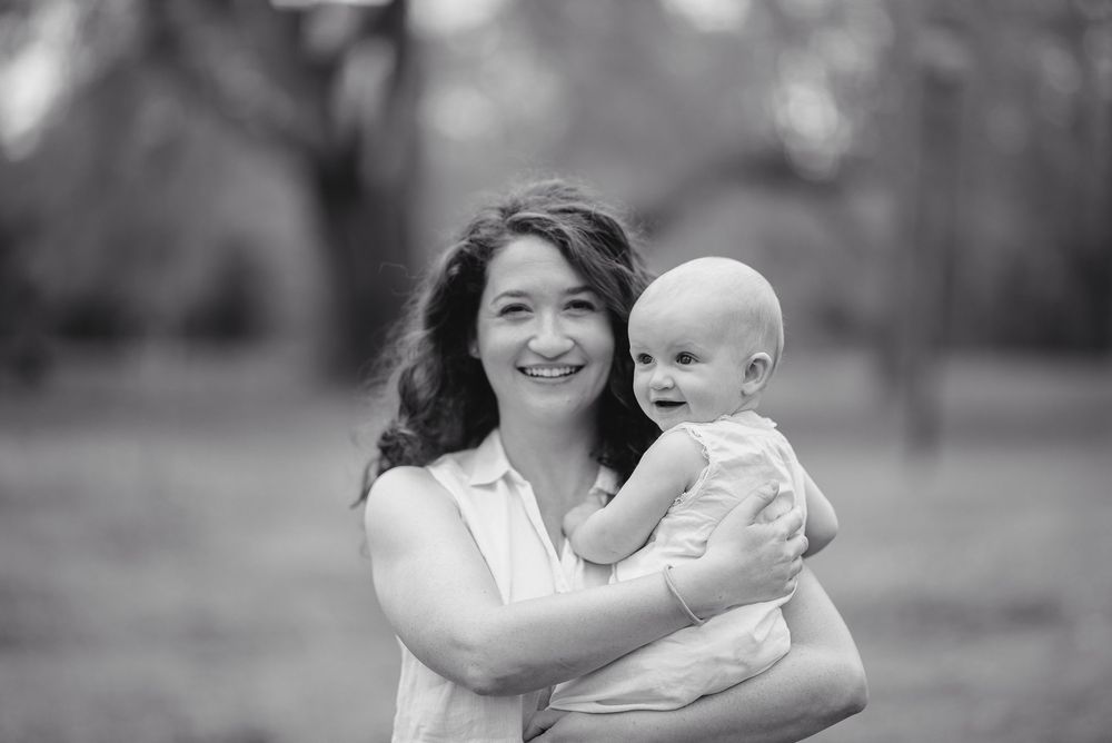 Black and white photo of a smiling woman holding a happy baby outdoors in a park setting.