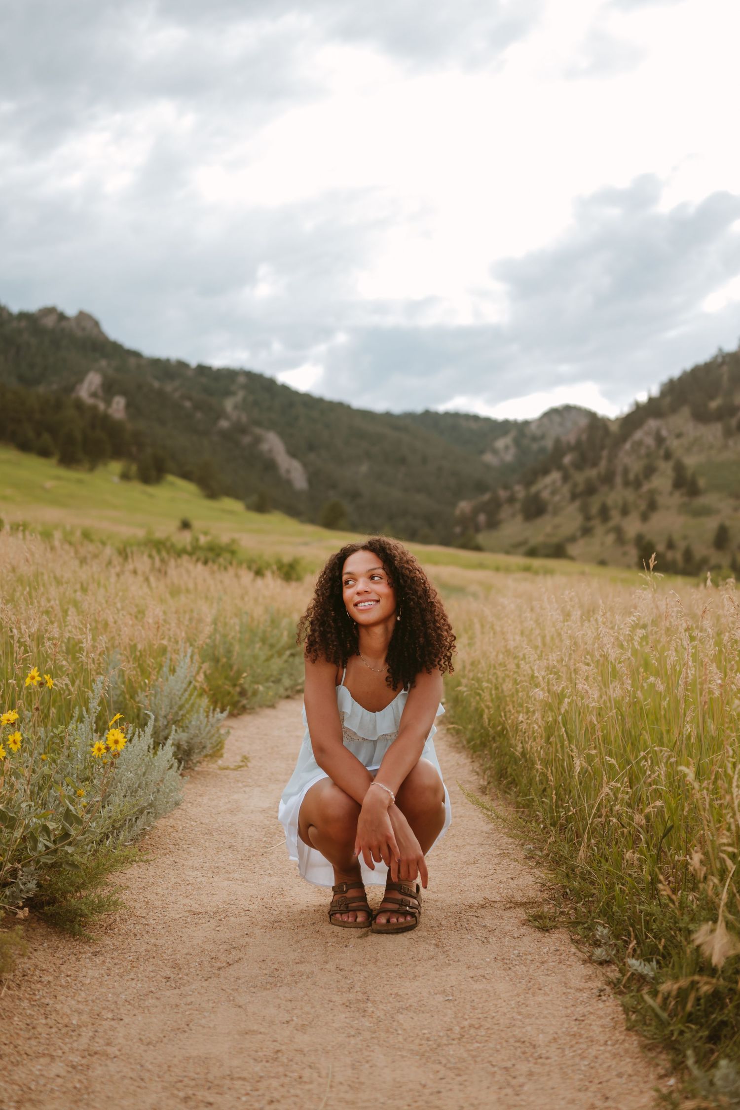 A person with long curly hair poses joyfully on a dirt hiking trail surrounded by tall grass and wildflowers in the mountains.