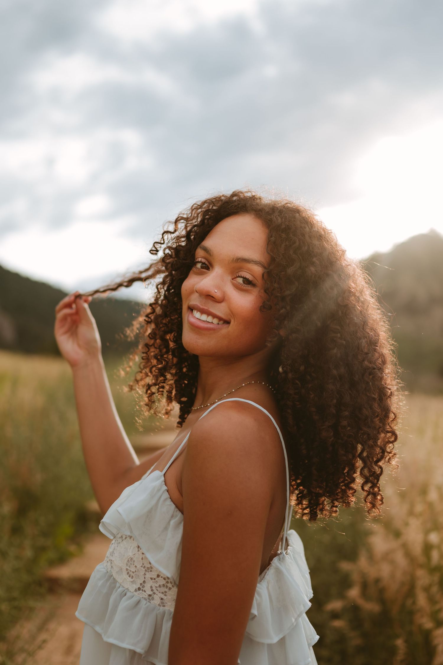 Golden sunlight illuminates a person with curly natural hair posing gracefully in a white ruffled top against nature's backdrop.