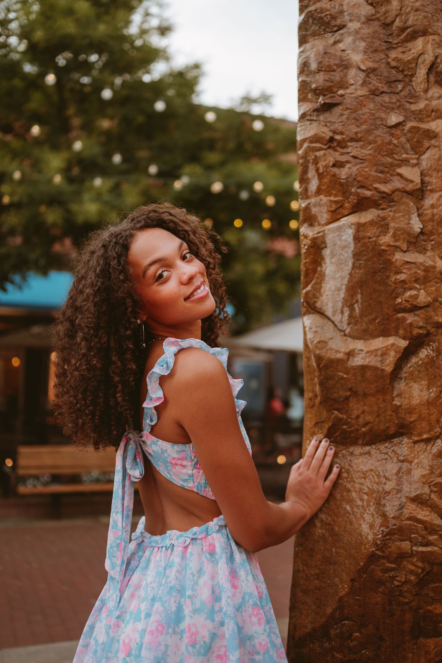 Happy young person in floral dress poses playfully against stone pillars during golden hour outdoor photoshoot.