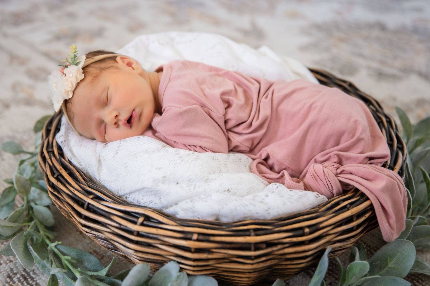 A sleeping newborn wearing a dusty pink sleeper rests in a wicker basket lined with white fabric and eucalyptus leaves.