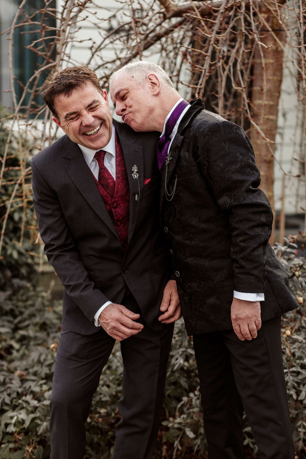 Groomsmen in black suits and burgundy ties share a candid laughing moment together outdoors.