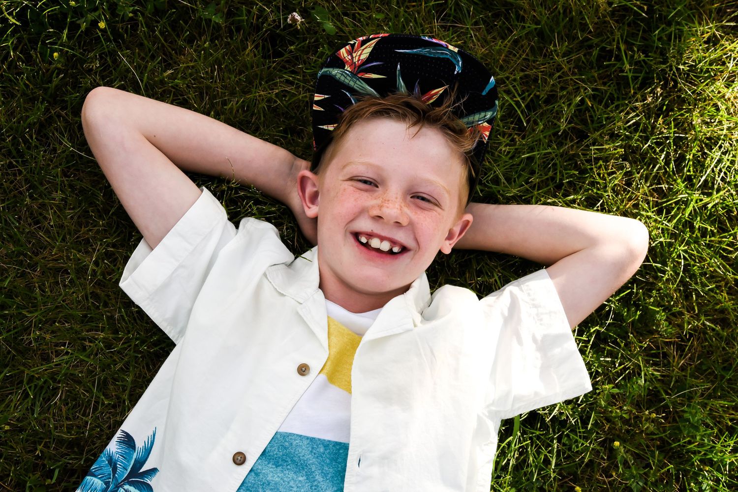 A smiling young boy with freckles is lying on his back in the grass, with his hands clasped behind his head. He is wearing a white and blue