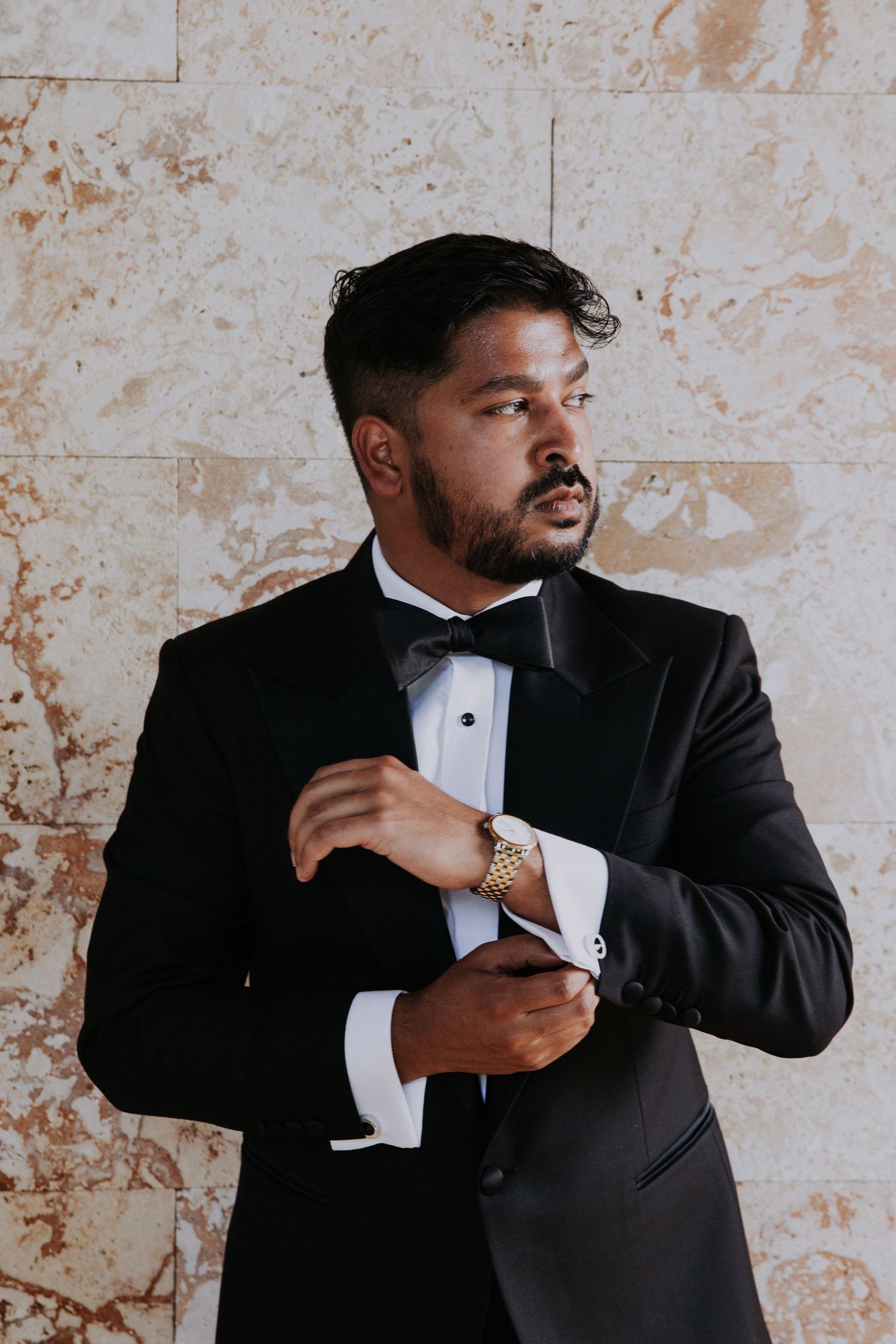 Groom adjusts cufflink while wearing black tuxedo against textured stone wall.