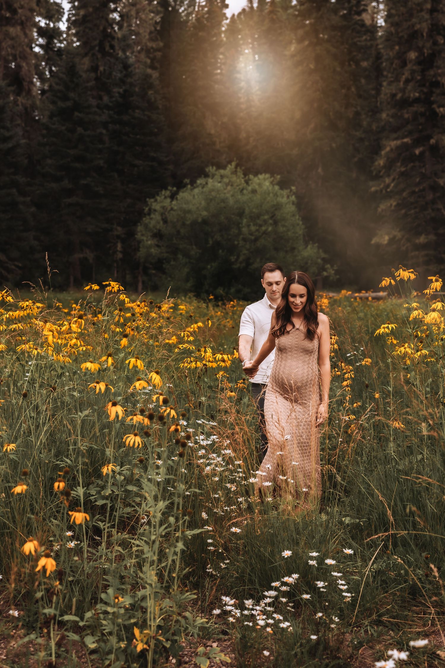 Pregnant couple walks through vibrant yellow wildflower field bathed in warm golden sunlight.