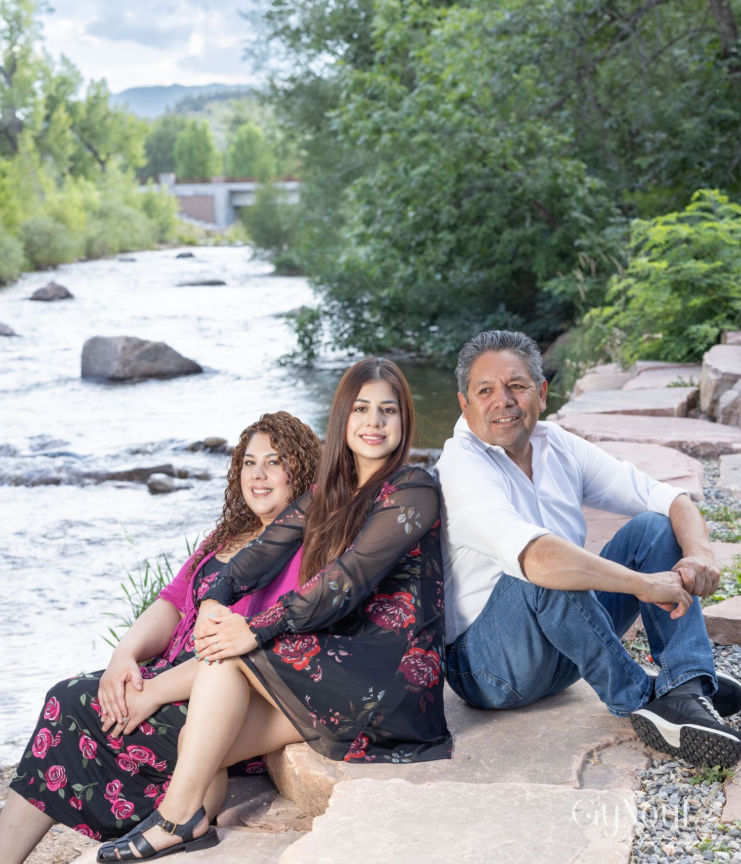 A group relaxes by a scenic riverside with trees and rocks, enjoying a peaceful moment in nature together.