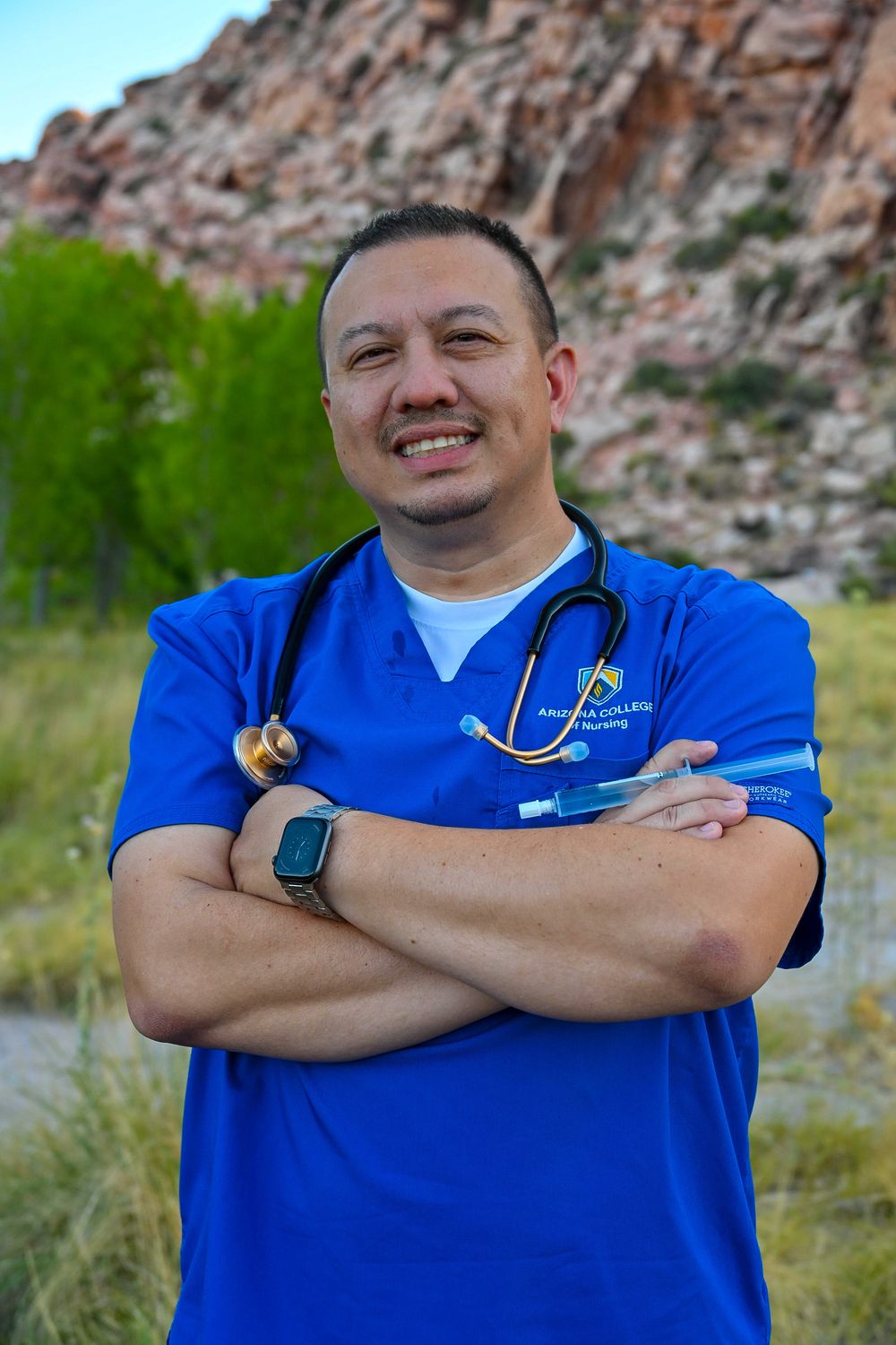 Smiling male nurse in blue scrubs with stethoscope standing arms crossed near rocky mountain landscape.