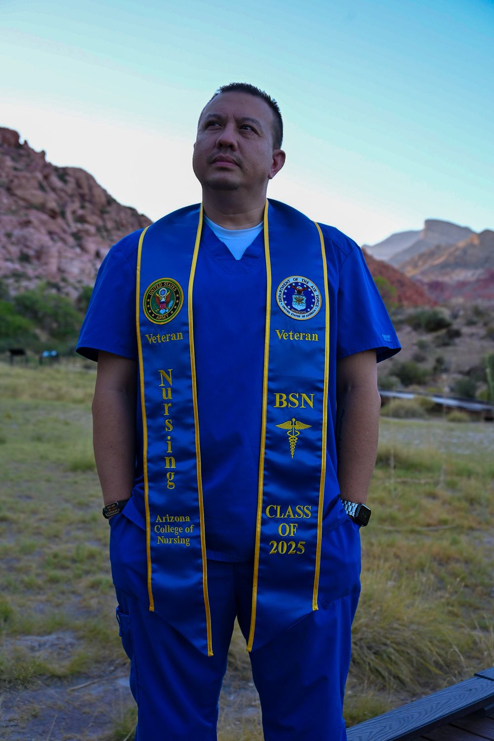 Male nursing BSN graduate in blue scrubs wearing veteran graduation stole reading Class of 2025 outdoors.