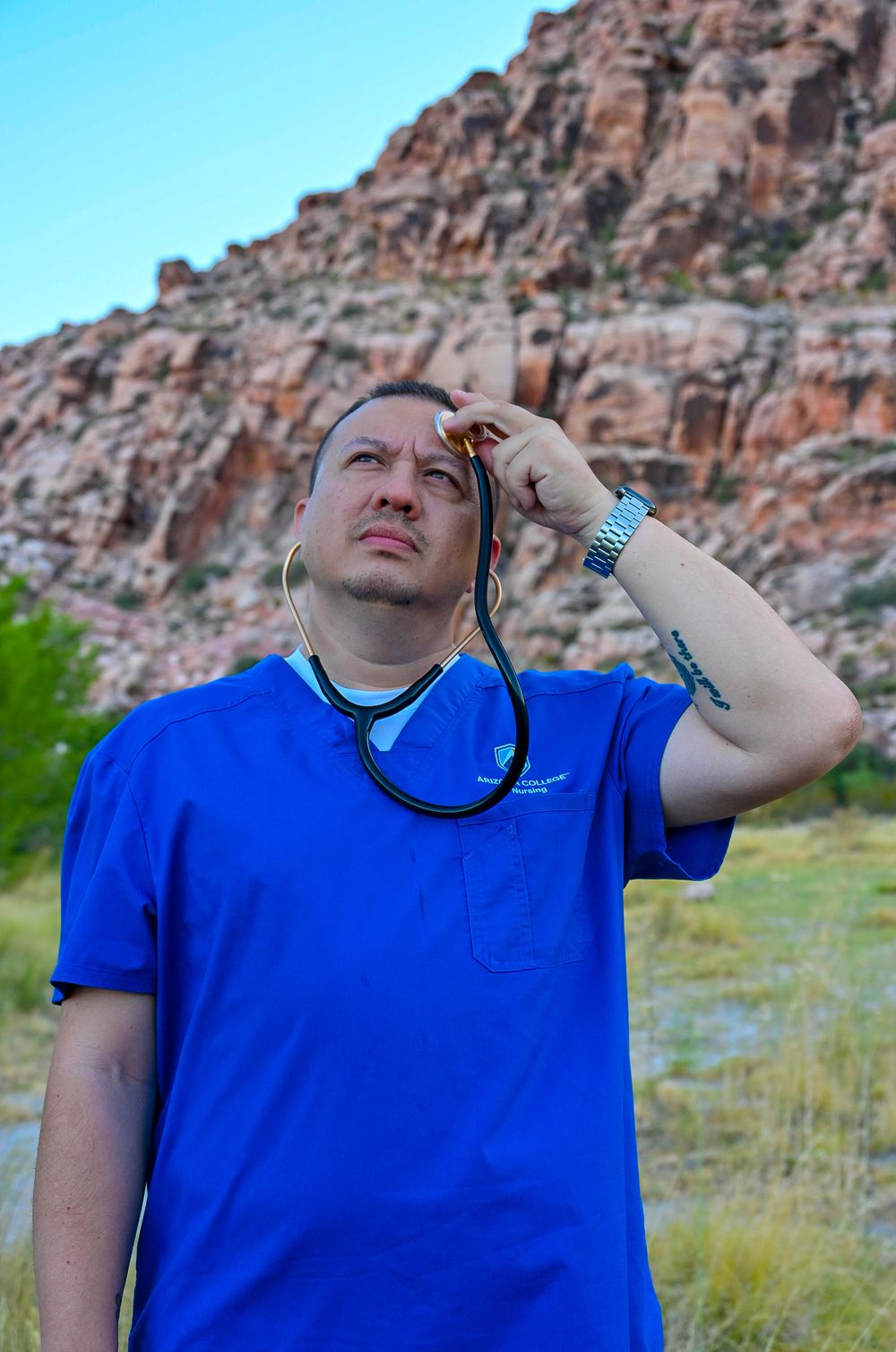Male nurse in blue scrubs holding stethoscope looking up against dramatic rocky mountain background.