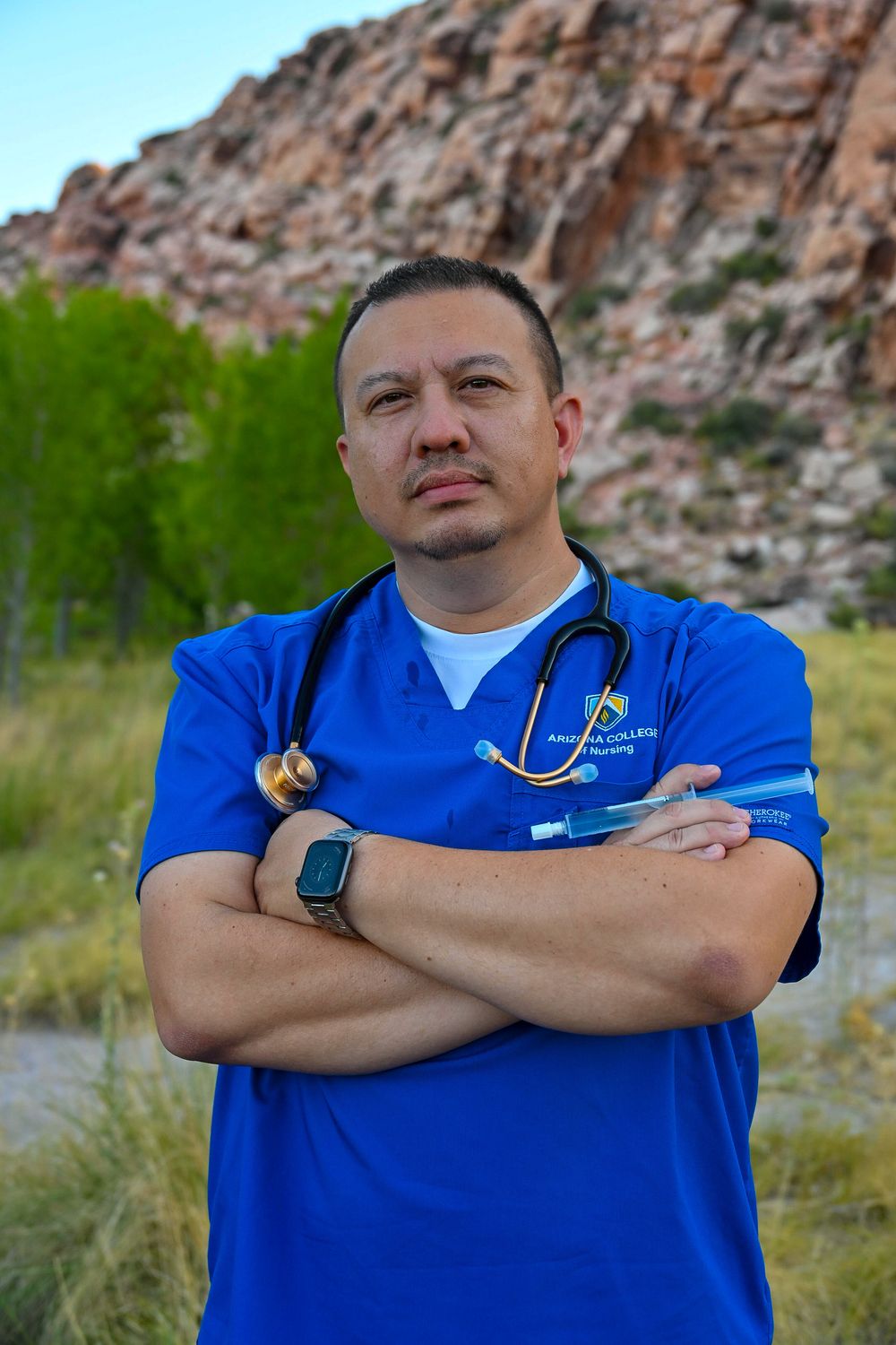 Nurse in blue scrubs with stethoscope stands arms crossed in front of rocky red canyon.