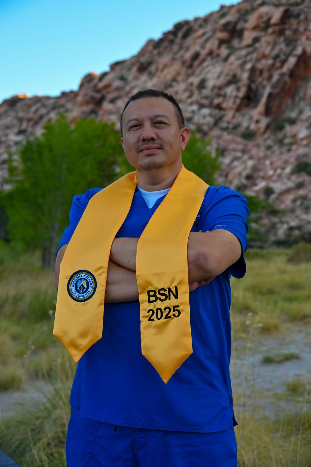 Man in blue scrubs with gold BSN 2025 graduation stole poses with arms crossed outdoors.
